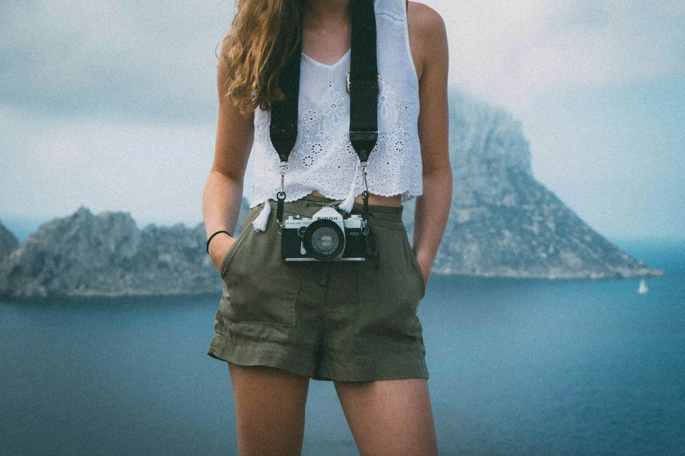 A woman wearing a white sleeveless eyelet top and olive green shorts, standing outdoors with a camera hanging around her neck, in front of a scenic mountainous landscape with water and trees.