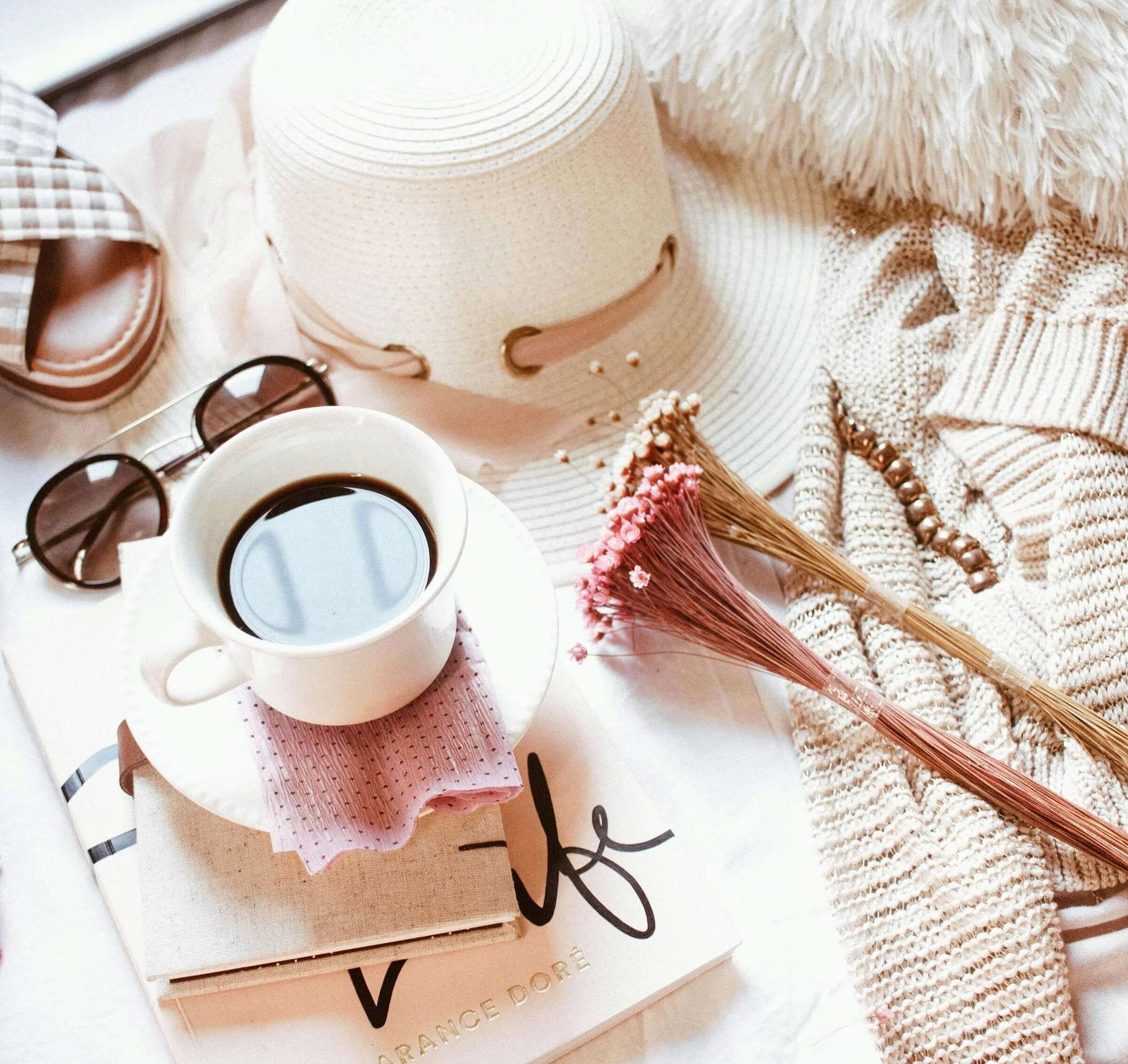 Close-up of a white sun hat, sunglasses, a cup of coffee, a pink and white stripy cloth, a beige book, a pearl necklace, and a bouquet of pink flowers, all arranged on a light-colored surface with some textured fabric.