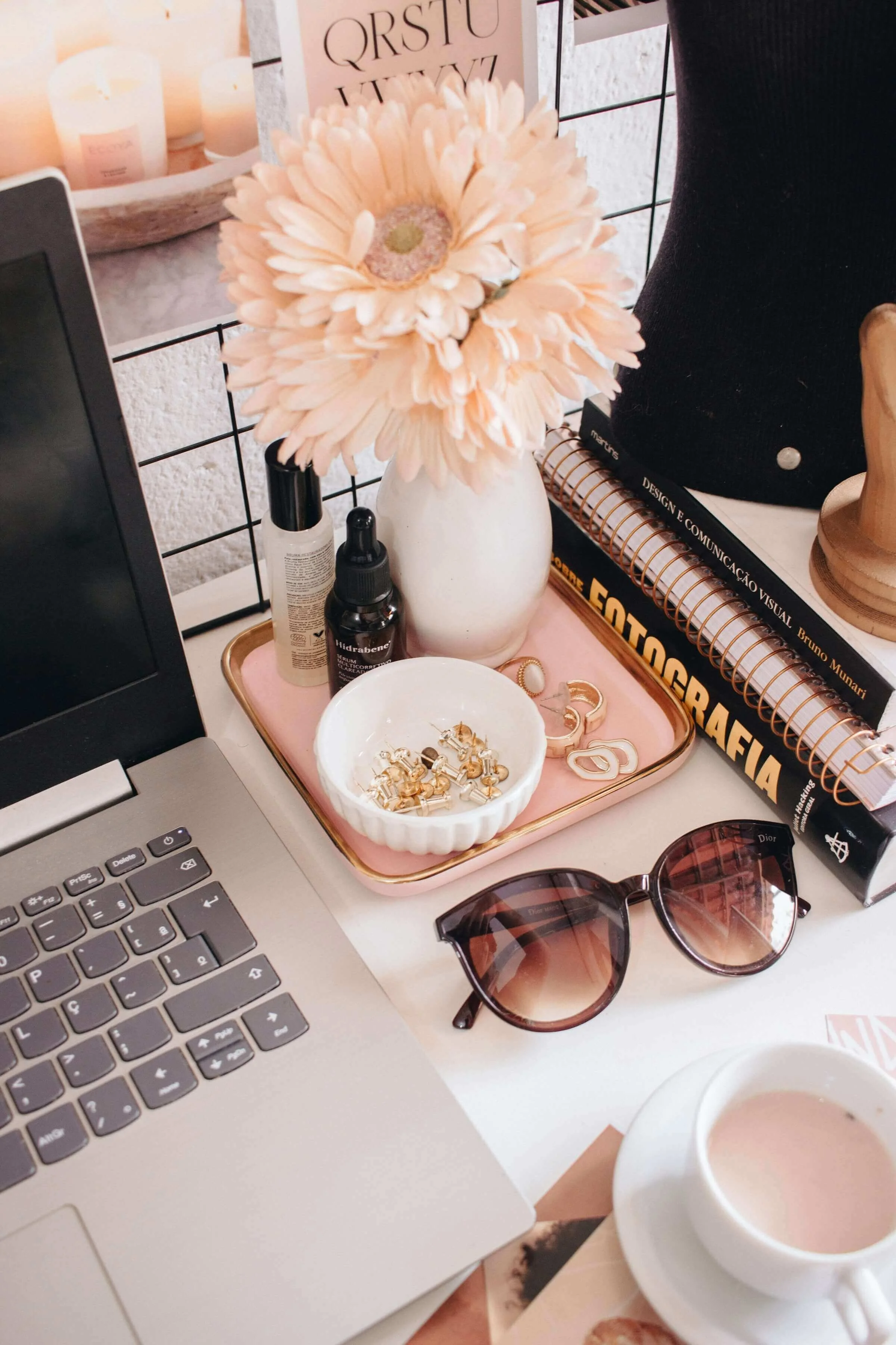 A workspace featuring a silver laptop with a black keyboard, a pink tray holding gold jewelry, a pair of sunglasses, a white cup with a hot beverage, decorative items, skincare products, books about design and typography, and a large beige vase with light pink flowers, all on a white surface.