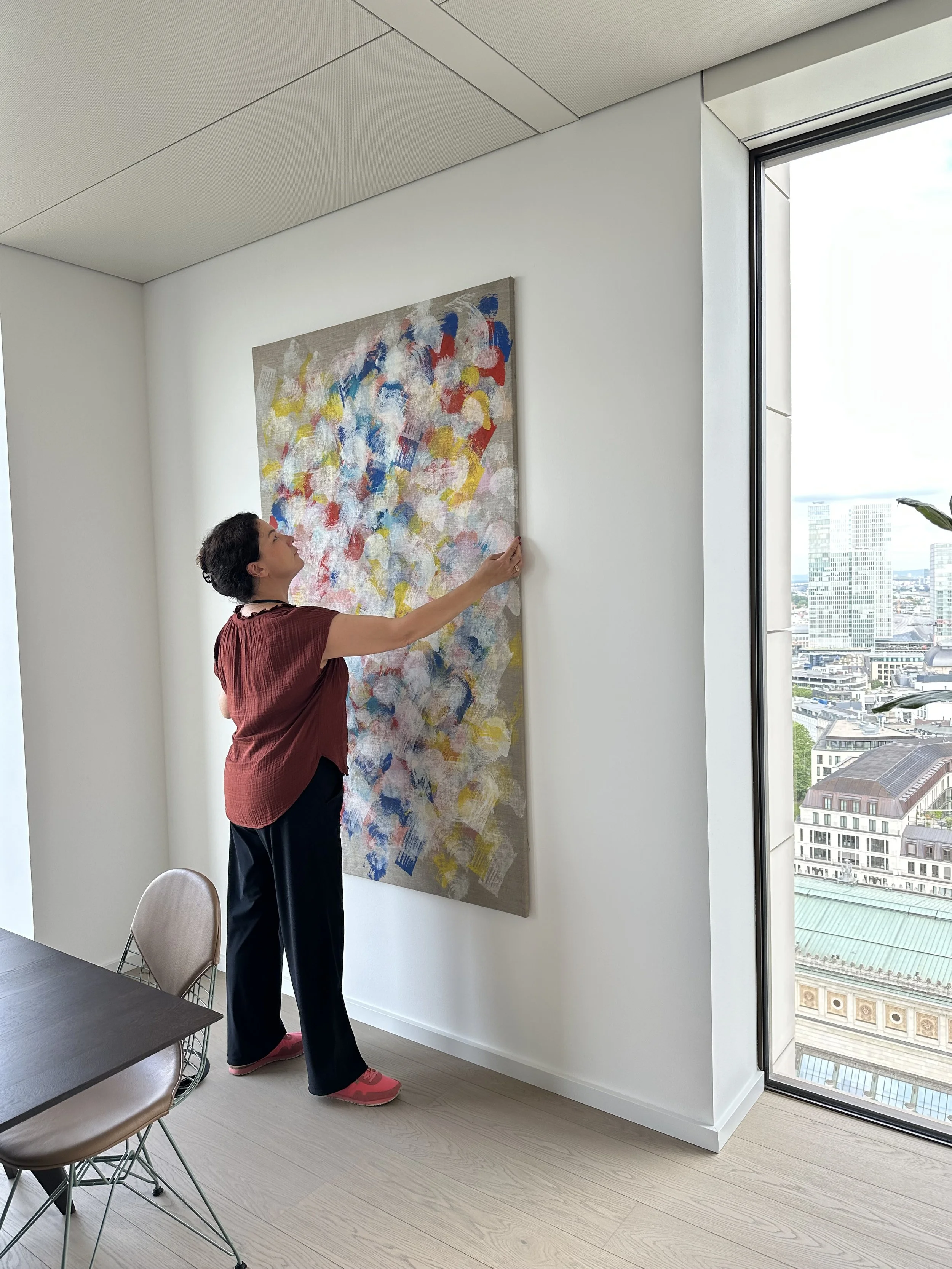 Frau hängt ein abstraktes Kunstwerk an die weiße Wand eines modernen Büros oder Apartments, mit großen Fenstern und Stadtblick.