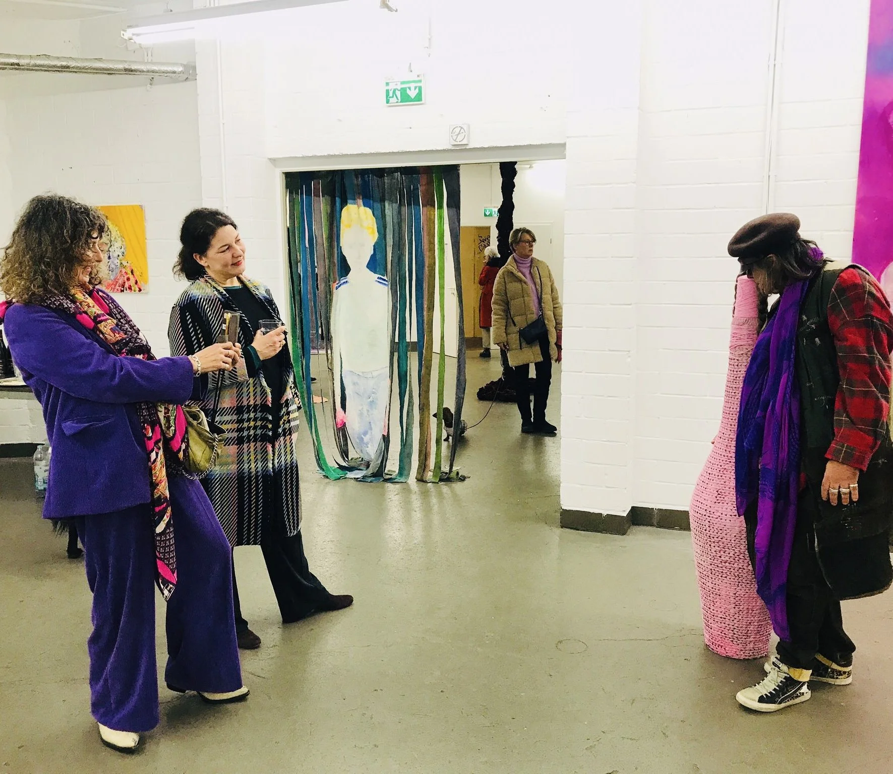 Menschen betrachten Kunstwerke in einer Galerie, darunter eine große bemalte Lichtinstallation und farbenfrohe Gemälde.