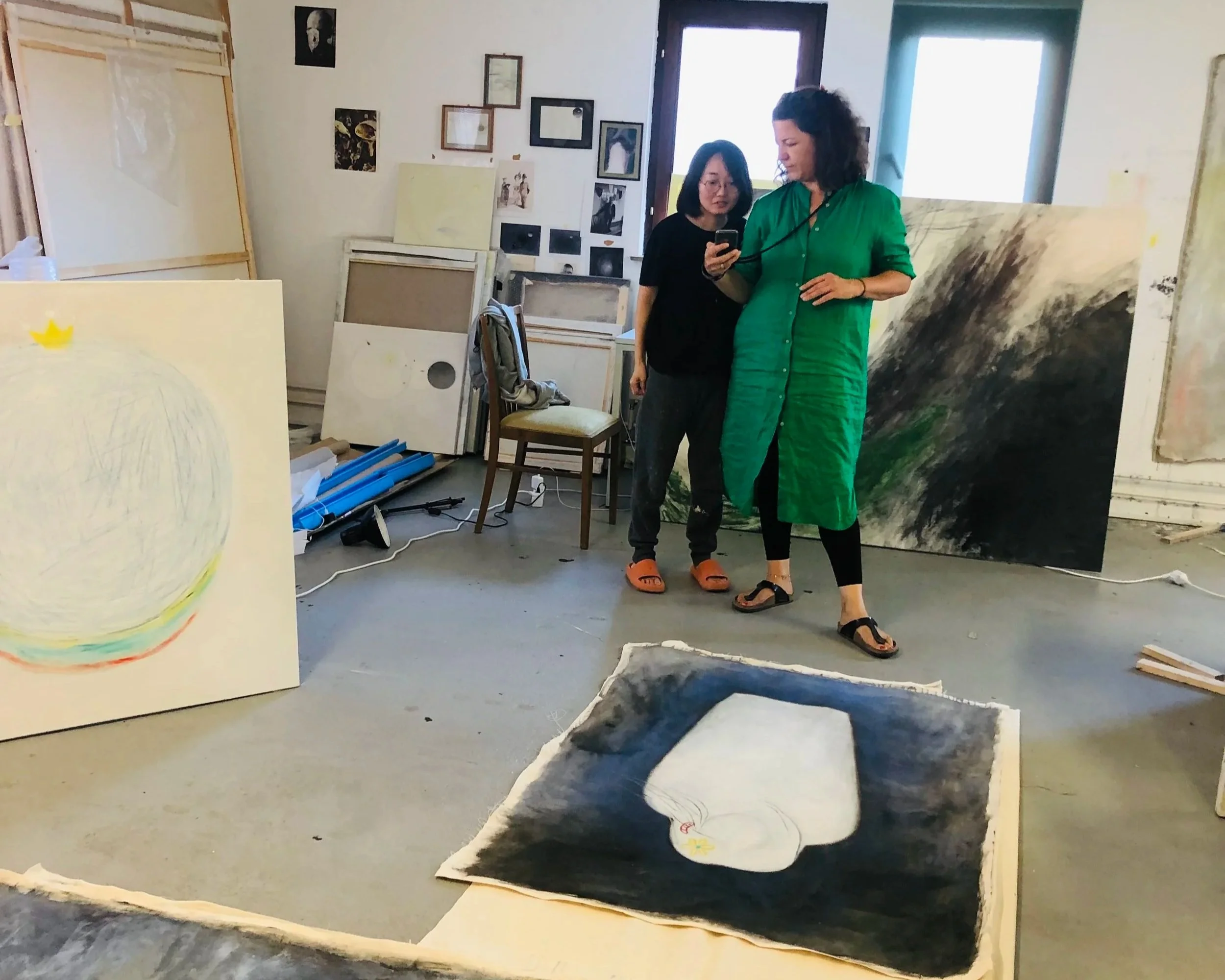 Zwei Frauen betrachten Kunstwerke in einem Kunstatelier, umgeben von Gemälden und Zeichenmaterialien.