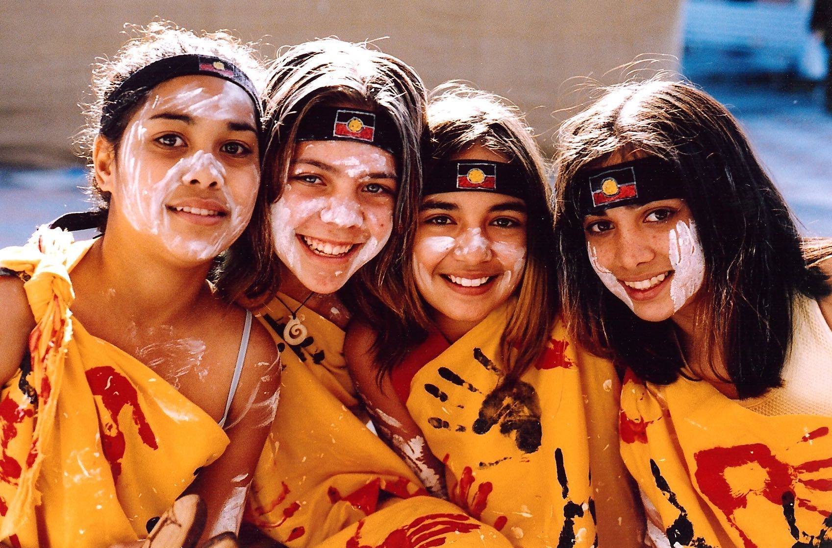 Four young girls smiling, wearing yellow shawls with red and black handprints and patterned headbands, and white paint on their faces.