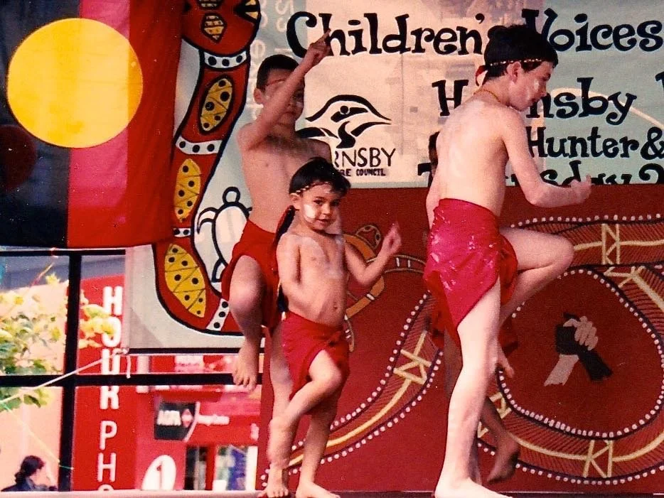 Children performing a traditional dance on stage at an event with cultural decorations and banners in the background.