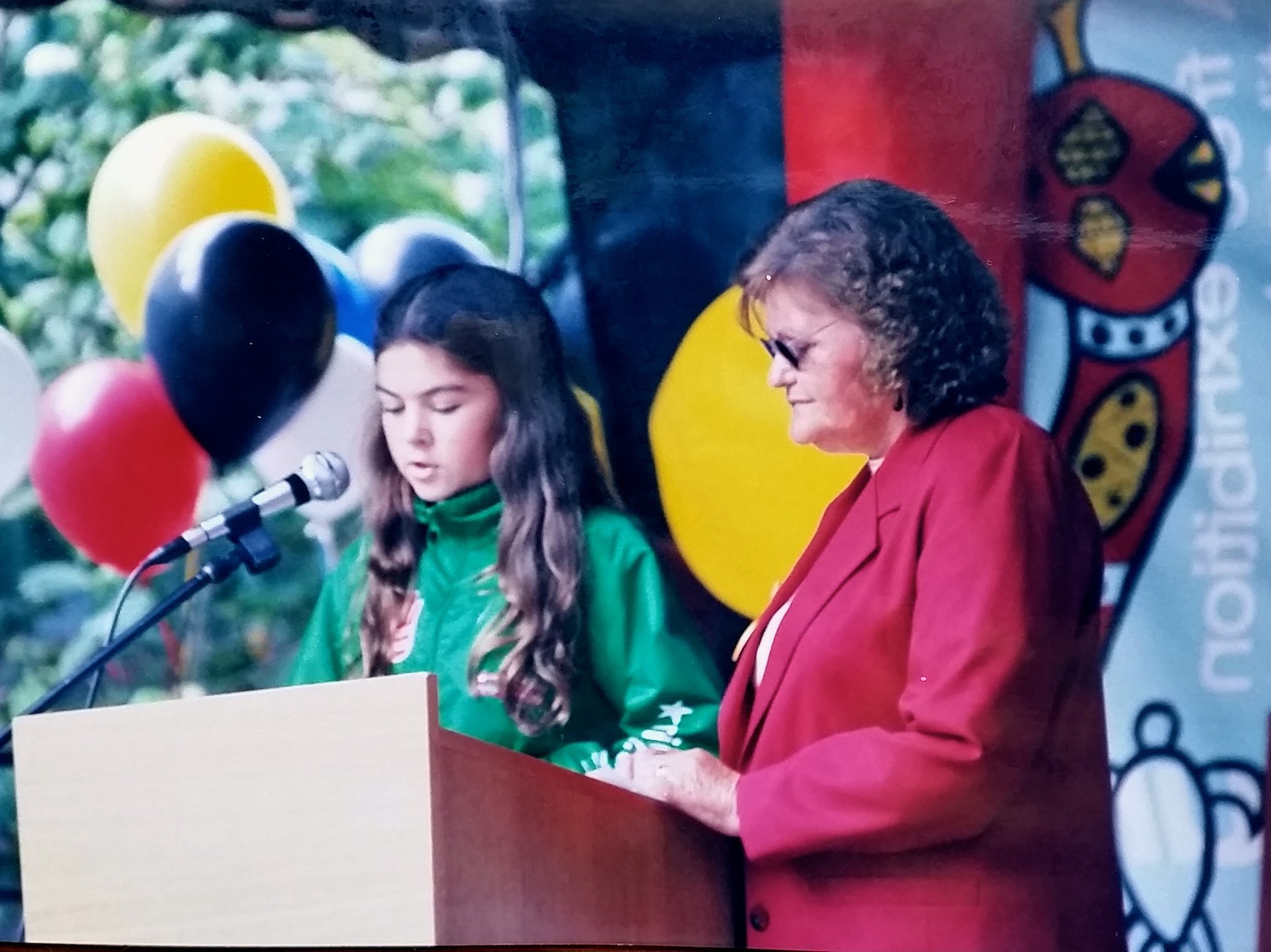 A young girl speaking at a podium with a microphone and an older woman standing beside her. They are outdoors, surrounded by colorful balloons and a backdrop with patterns and text. The girl has long hair and is wearing a green jacket, while the woma