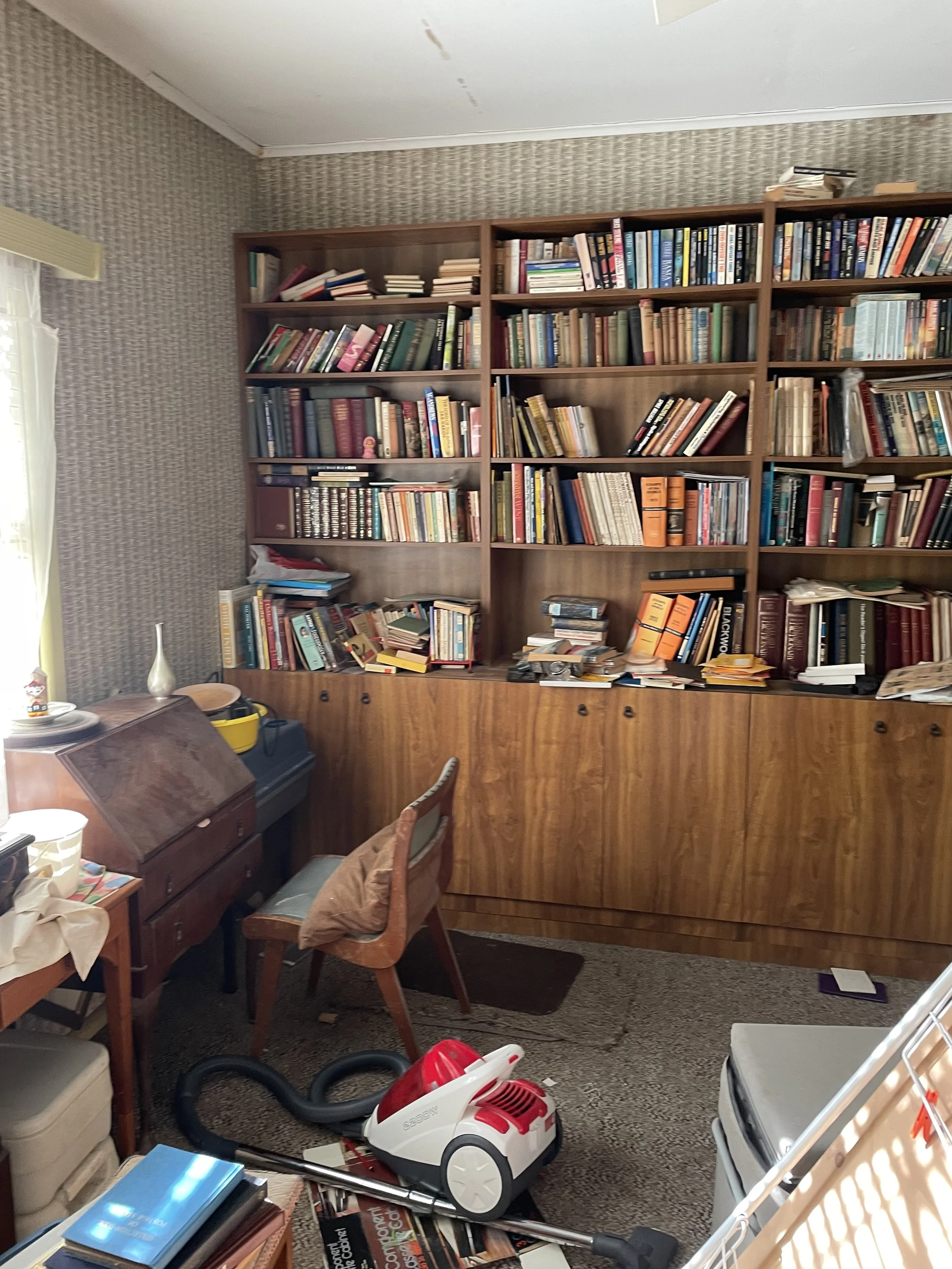 A cluttered room with a large wooden bookshelf filled with books, a small desk with a chair, and a vacuum cleaner on the carpet.