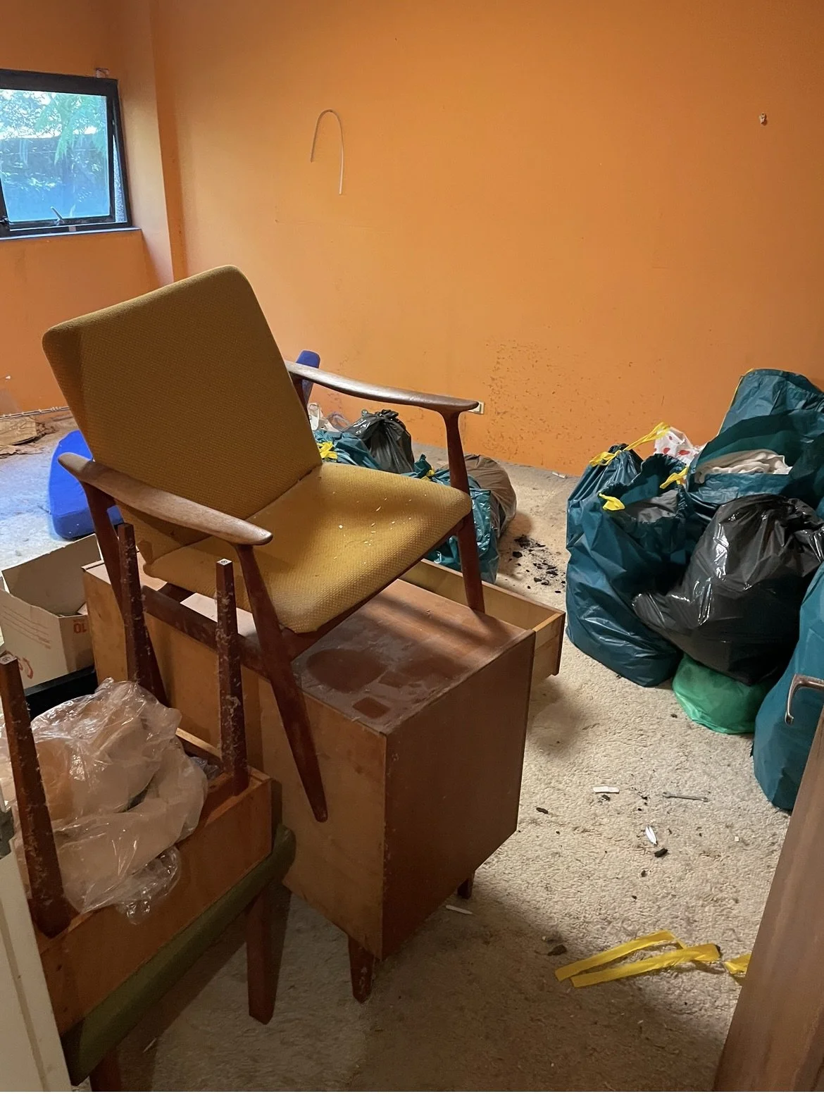 Room filled with trash bags, an old yellow armchair on top of a wooden cabinet, and scattered debris with an orange painted wall and a small window.