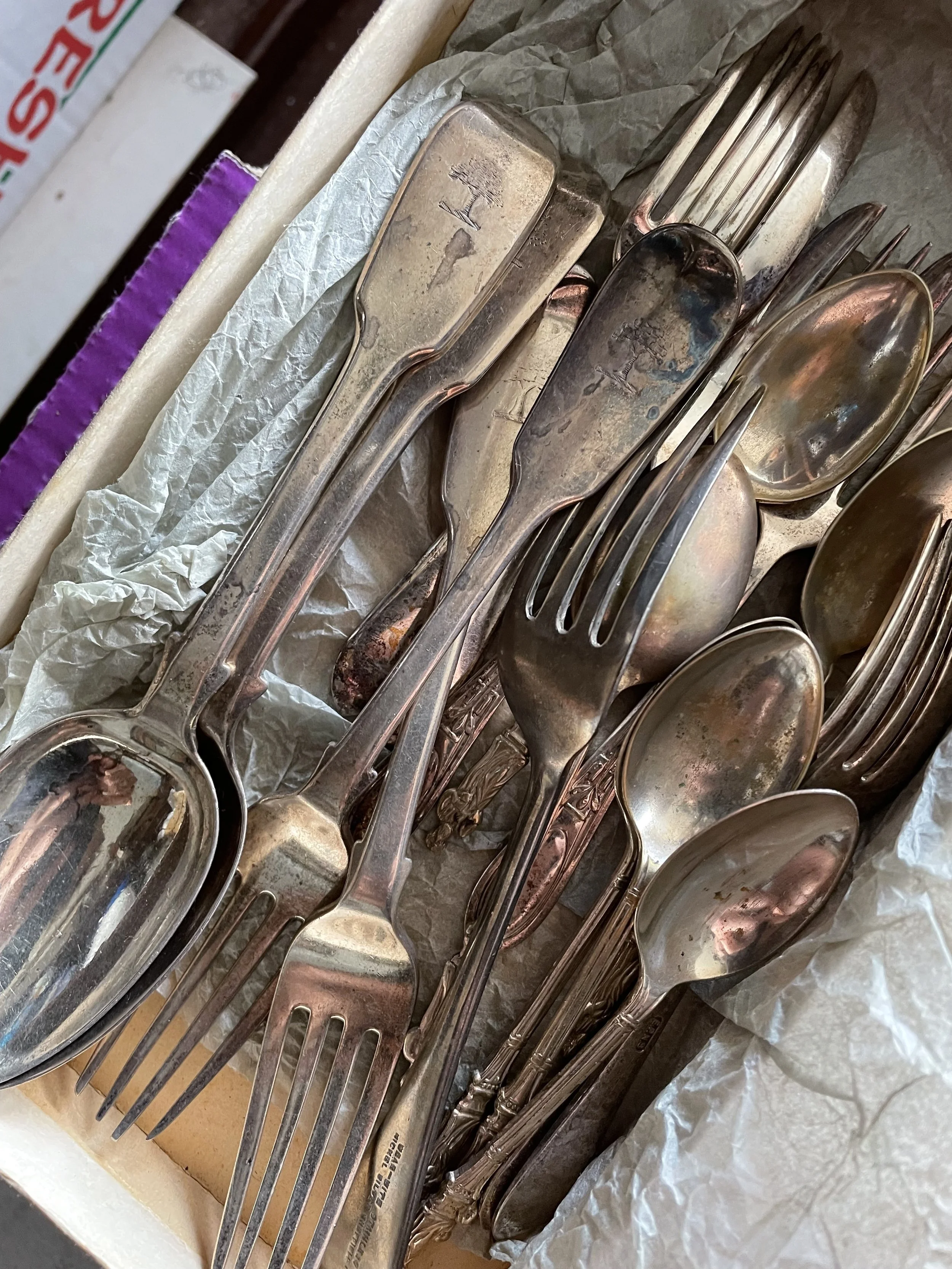 A collection of old, tarnished silverware including forks and spoons, placed on crinkled paper inside a box.