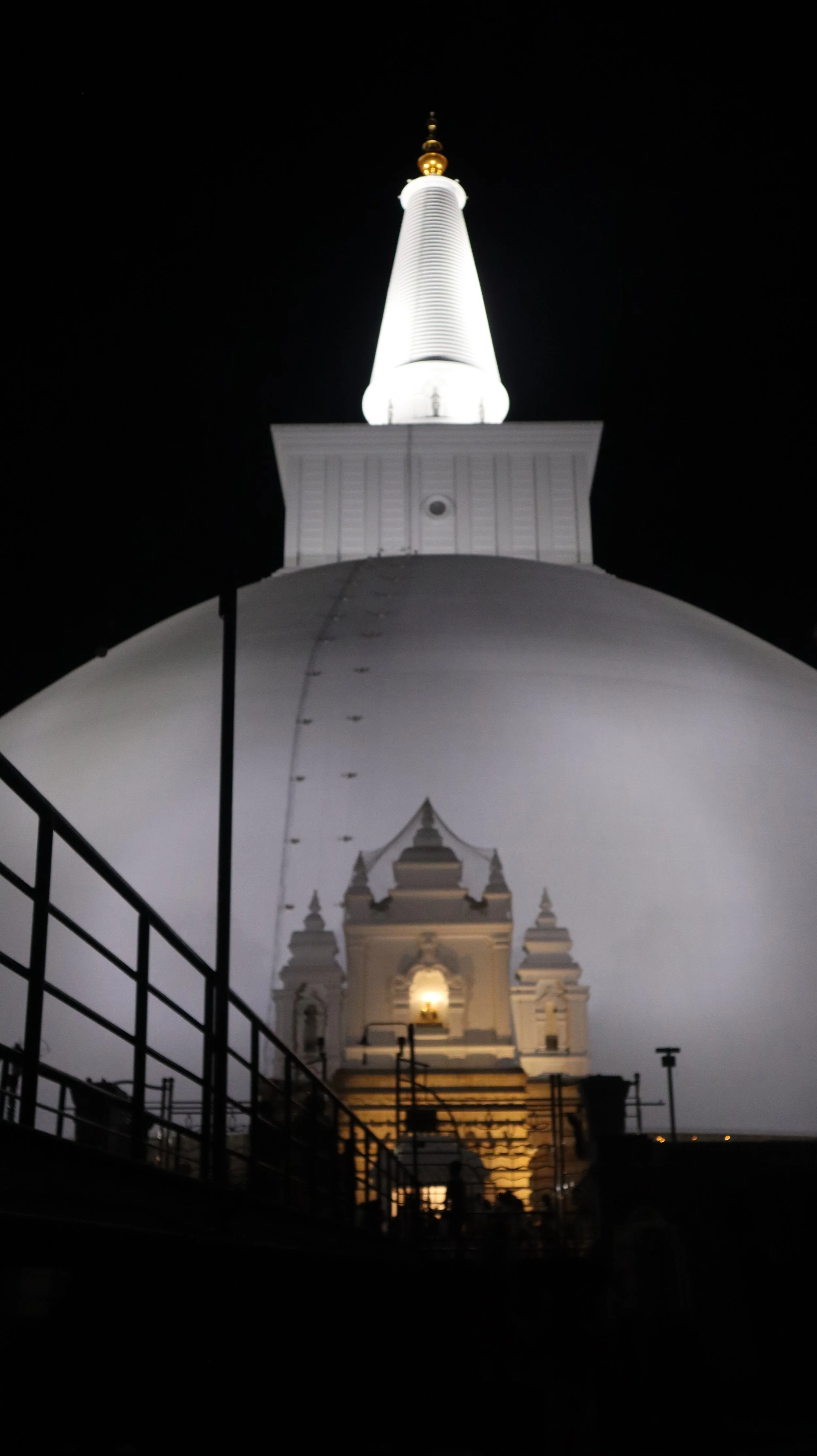 Nighttime photograph of a white Buddhist stupa with a spire, illuminated from below, against a dark sky.
