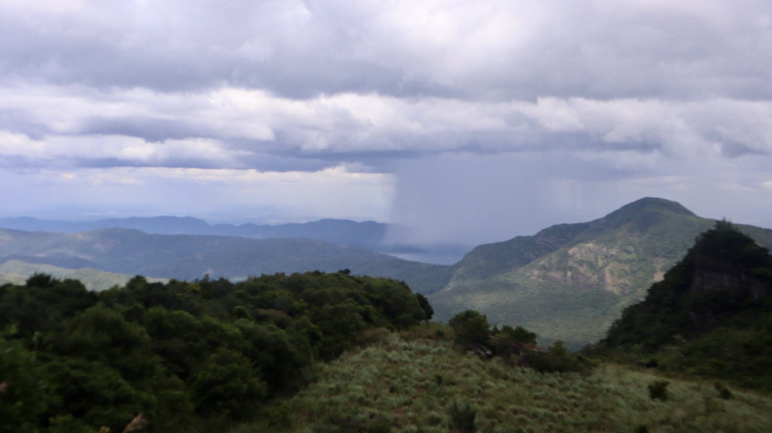 Scenic view of green mountains under cloudy sky with rain in the distance.