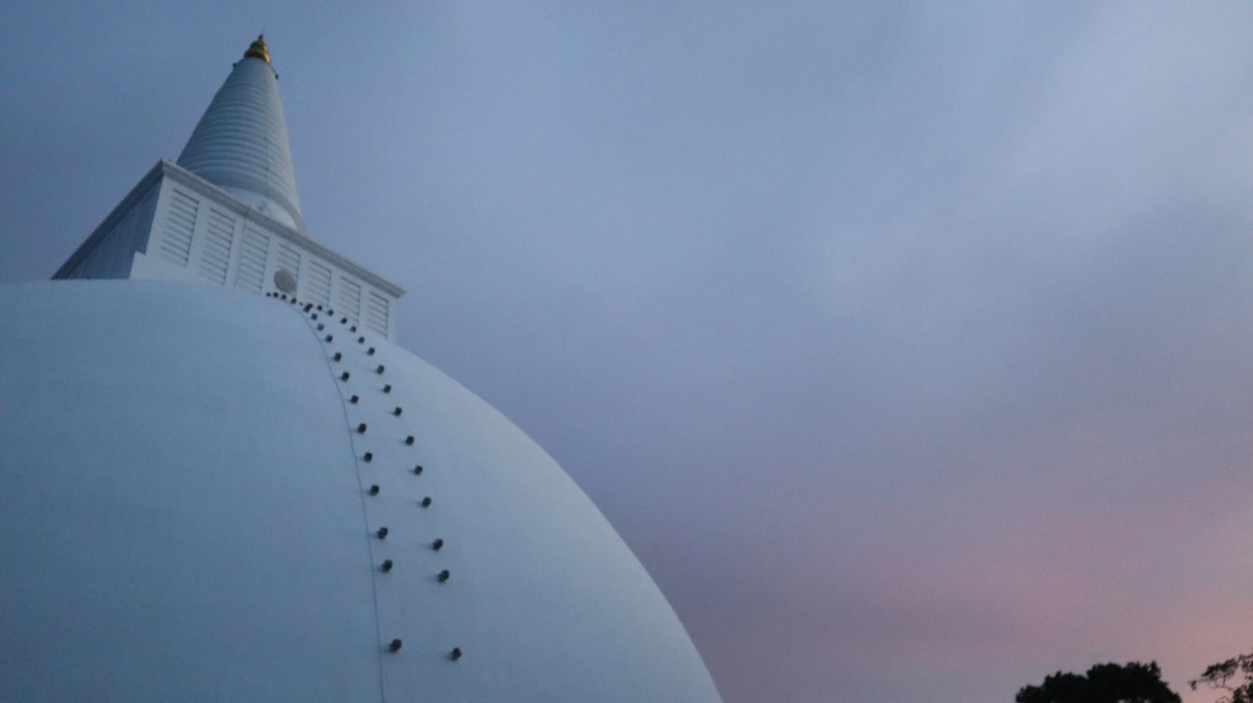 Low-angle view of a white stupa or pagoda with a golden tip, set against a dusky sky with a hint of sunset colors, and a silhouette of trees in the background.