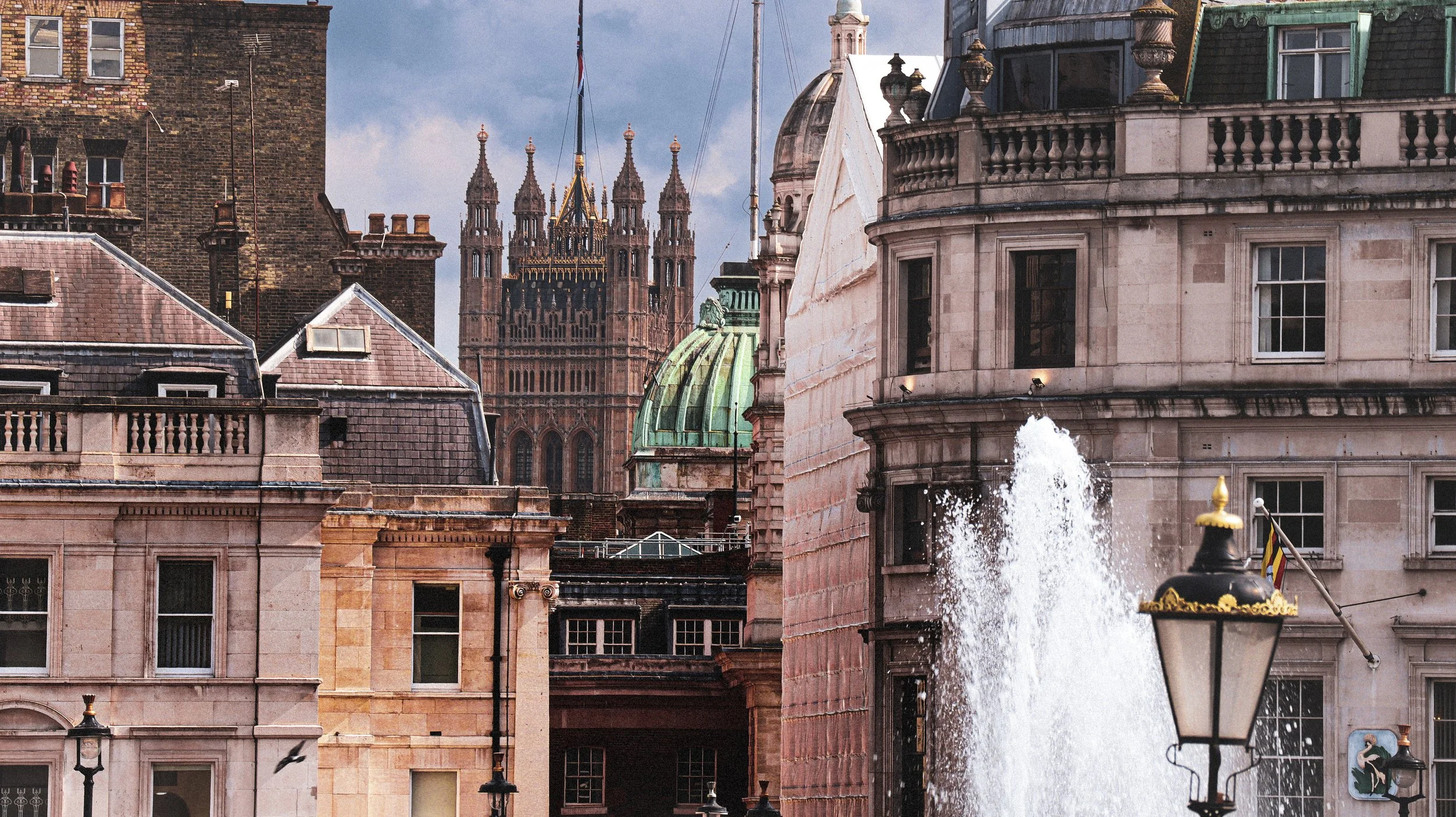 A cityscape featuring historic buildings and a fountain in the foreground, with a large gothic-style church or cathedral with spires in the background.