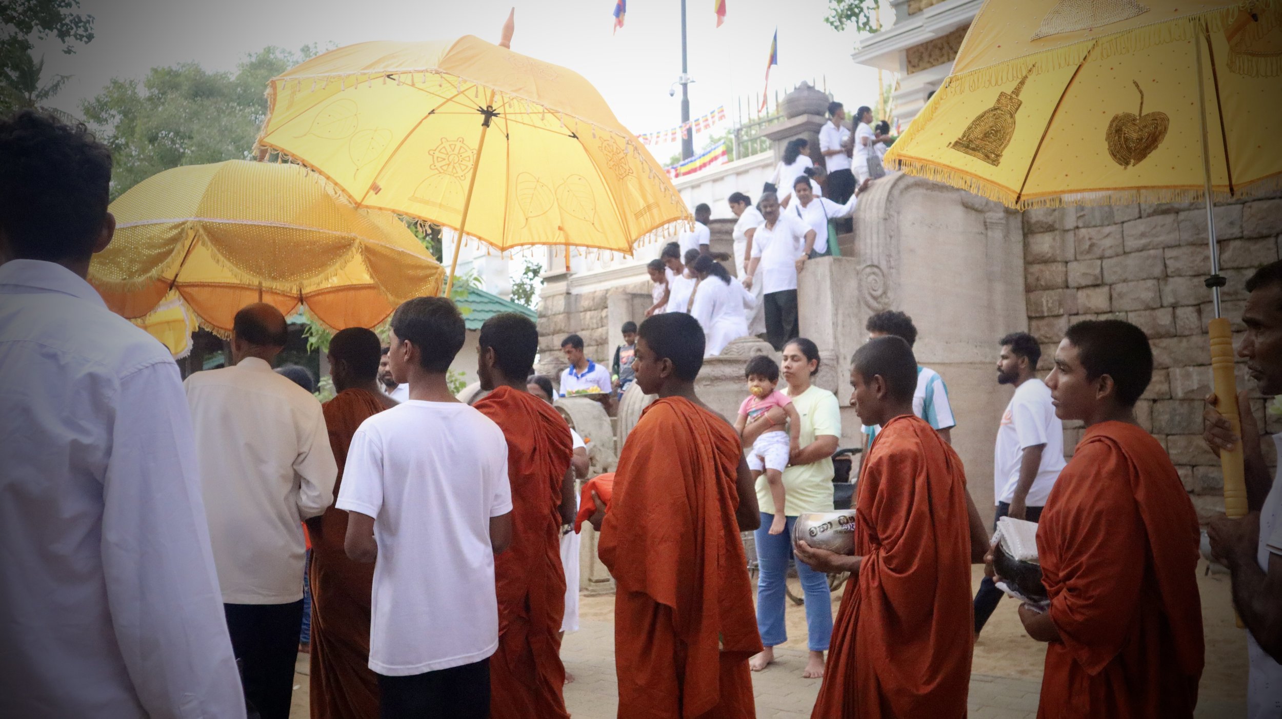 People participating in a religious or cultural procession, with some individuals wearing orange robes, holding umbrellas, and carrying offerings, in front of a temple or historical site with stone steps and flags.