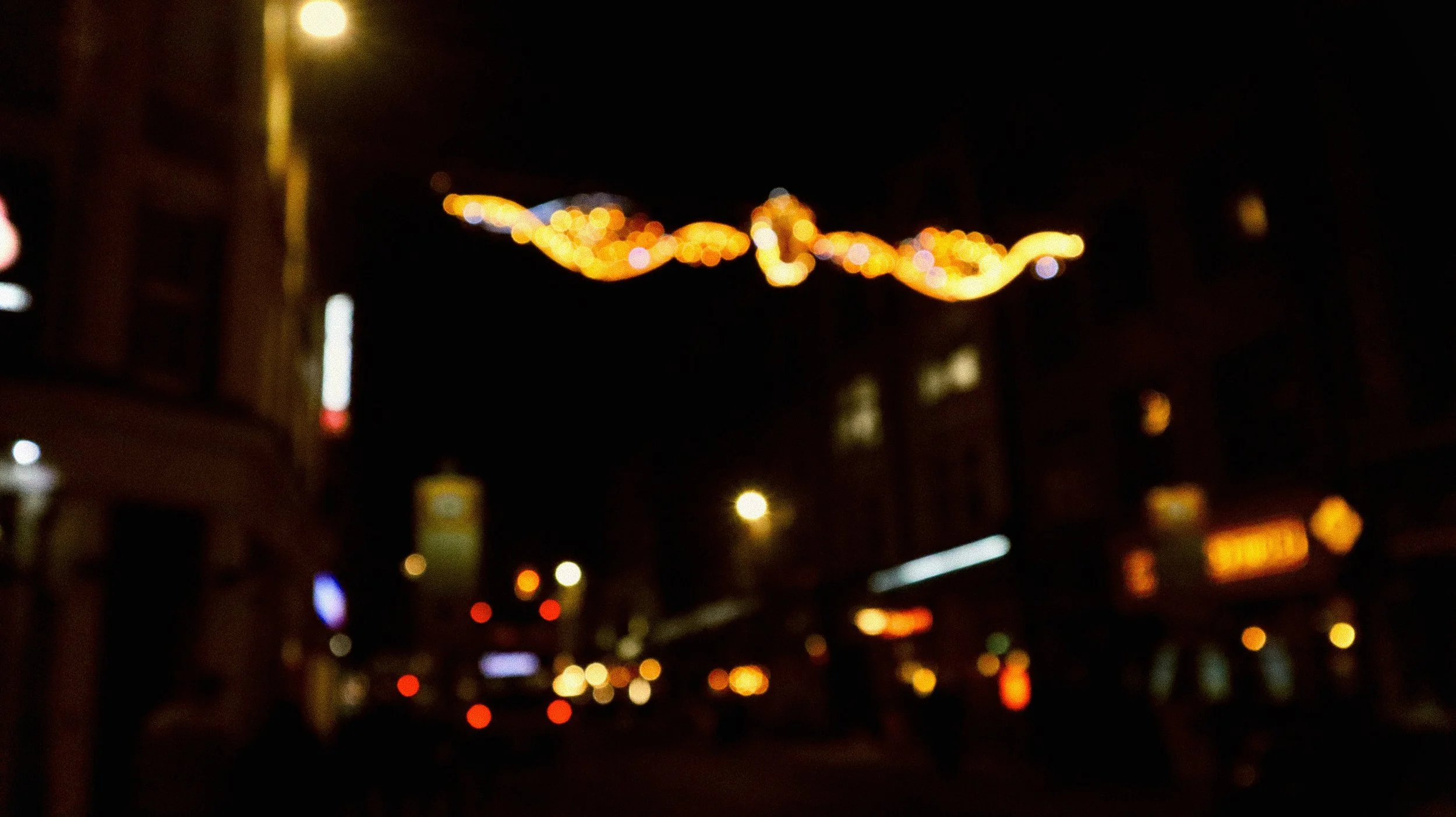 Blurred city street at night with colorful string lights hanging across the sky, and various illuminated signs and streetlights