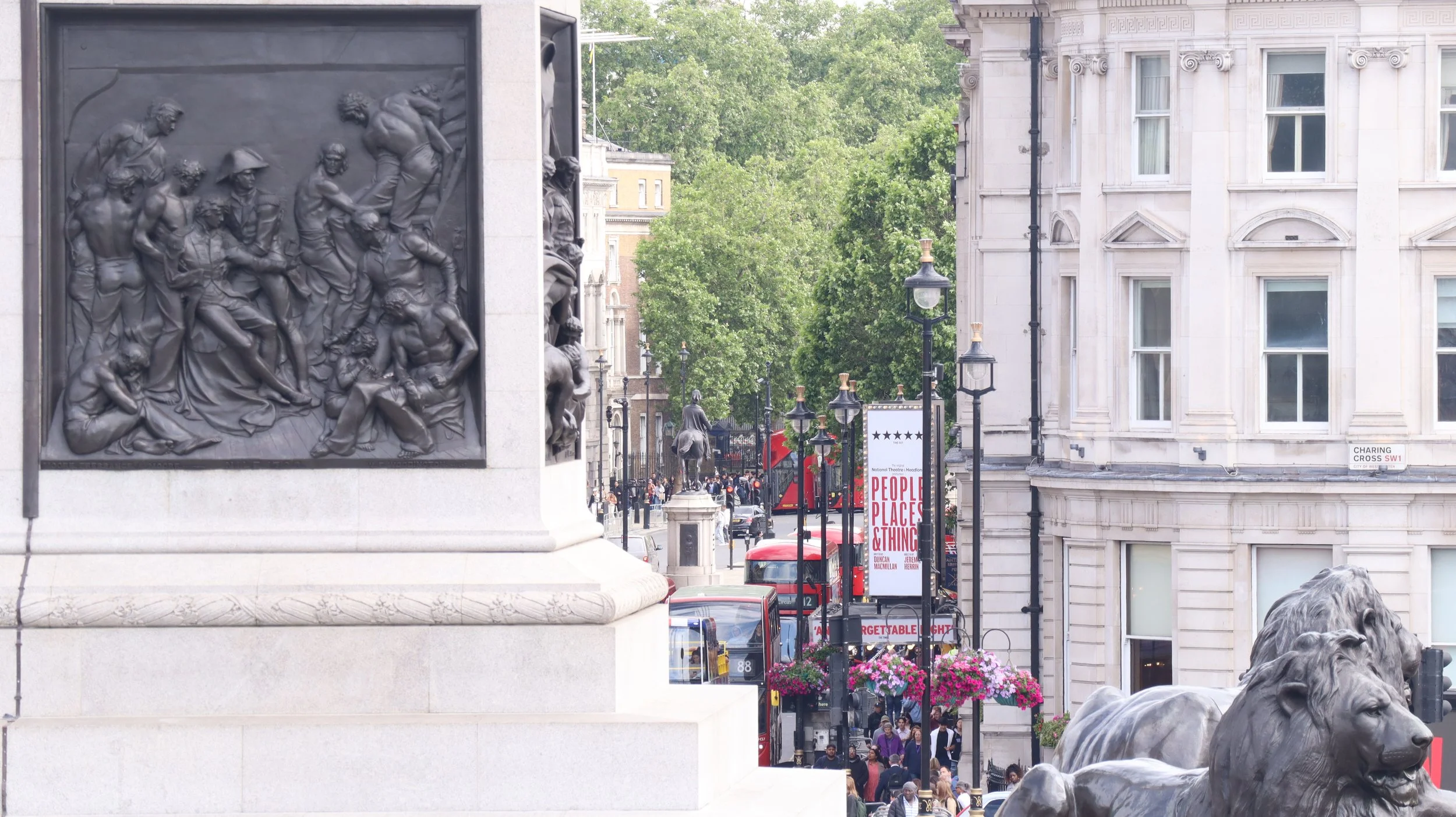 Street scene with historical monuments, street lamps, people, and red buses in London, with a large stone monument with a bronze bas-relief sculpture on the left and bronze lion statues at the bottom right.