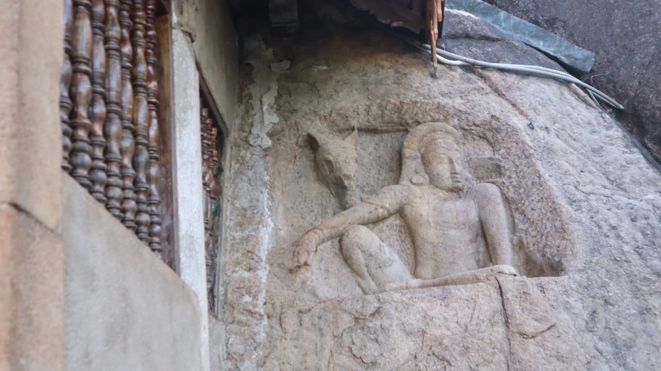 Ancient carved stone sculpture of a figure with a headdress, sitting on a flat surface, embedded in a stone wall, next to some electrical wires.