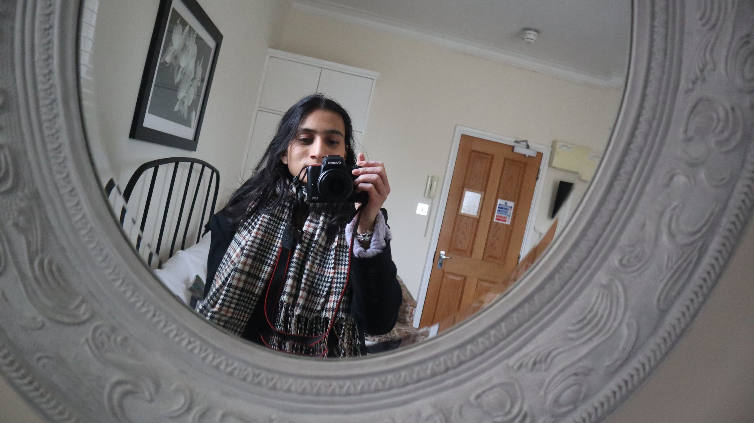 A woman taking a selfie in a mirror with a camera, visible in a bedroom with a wooden door, framed artwork, and striped headboard.