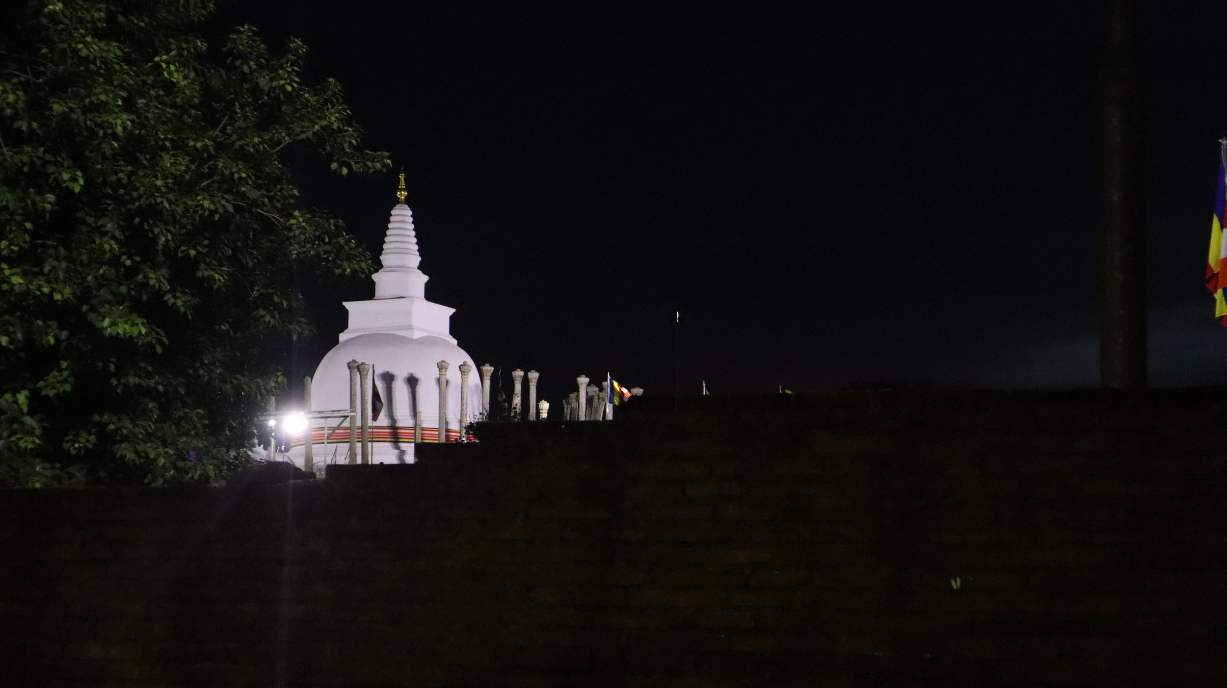 Nighttime view of a white stupa with a gold finial, surrounded by columns and flags, partially obscured by a dark brick wall in the foreground.
