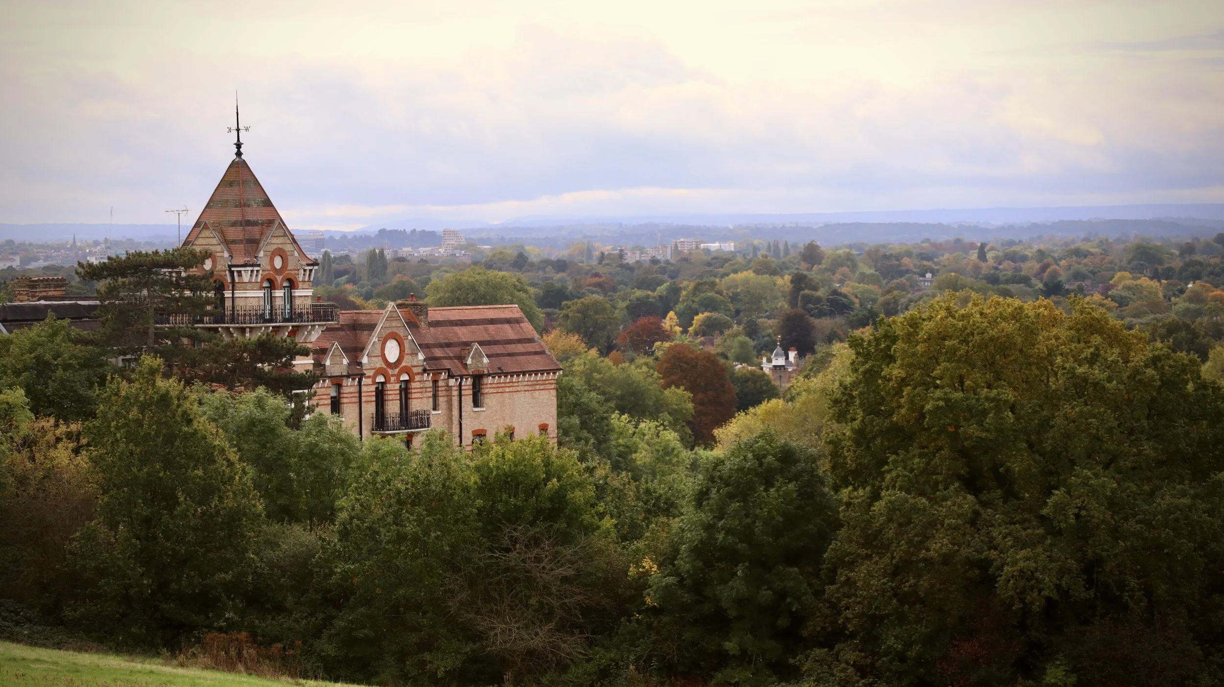 A historic brick building with a tall tower and red-tiled roofs, surrounded by lush green trees, overlooking a landscape with more trees and distant cityscape under a cloudy sky.