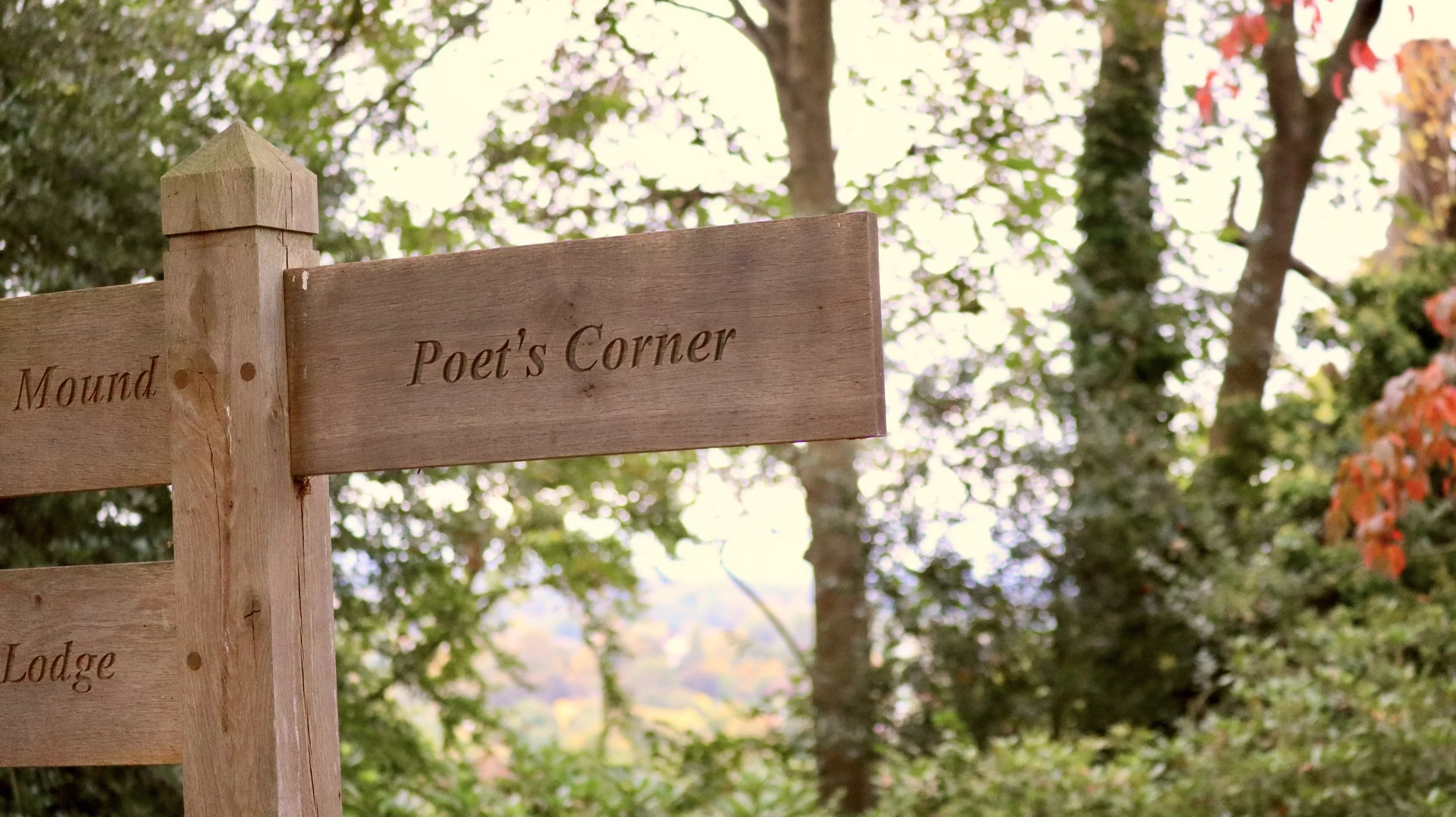 Wooden signpost with the words "Poet's Corner" engraved, surrounded by trees with green and orange leaves in the background.