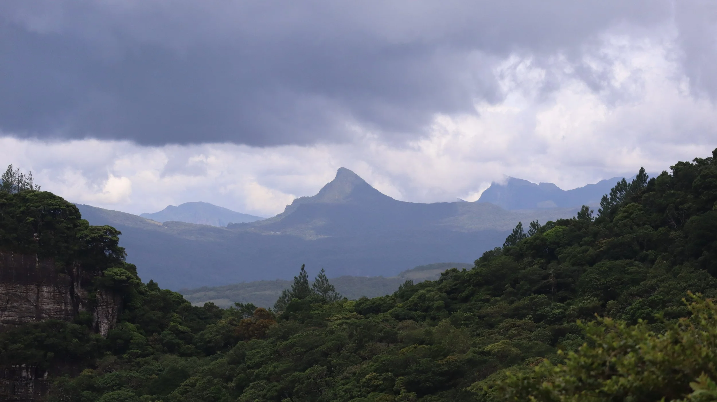 Mountain range with cloud-covered sky and lush green forested hills in the foreground.