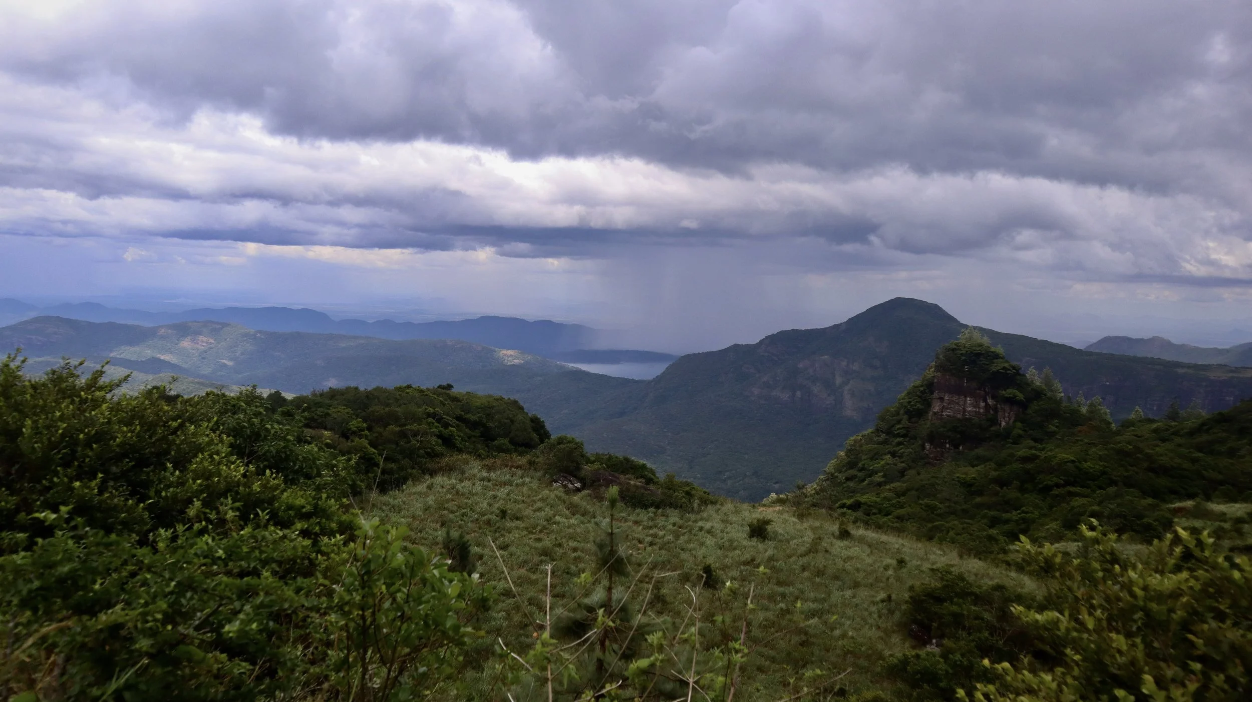 Landscape view of green mountains under dark, cloudy sky with rain in the distance and a lake visible between the mountains.