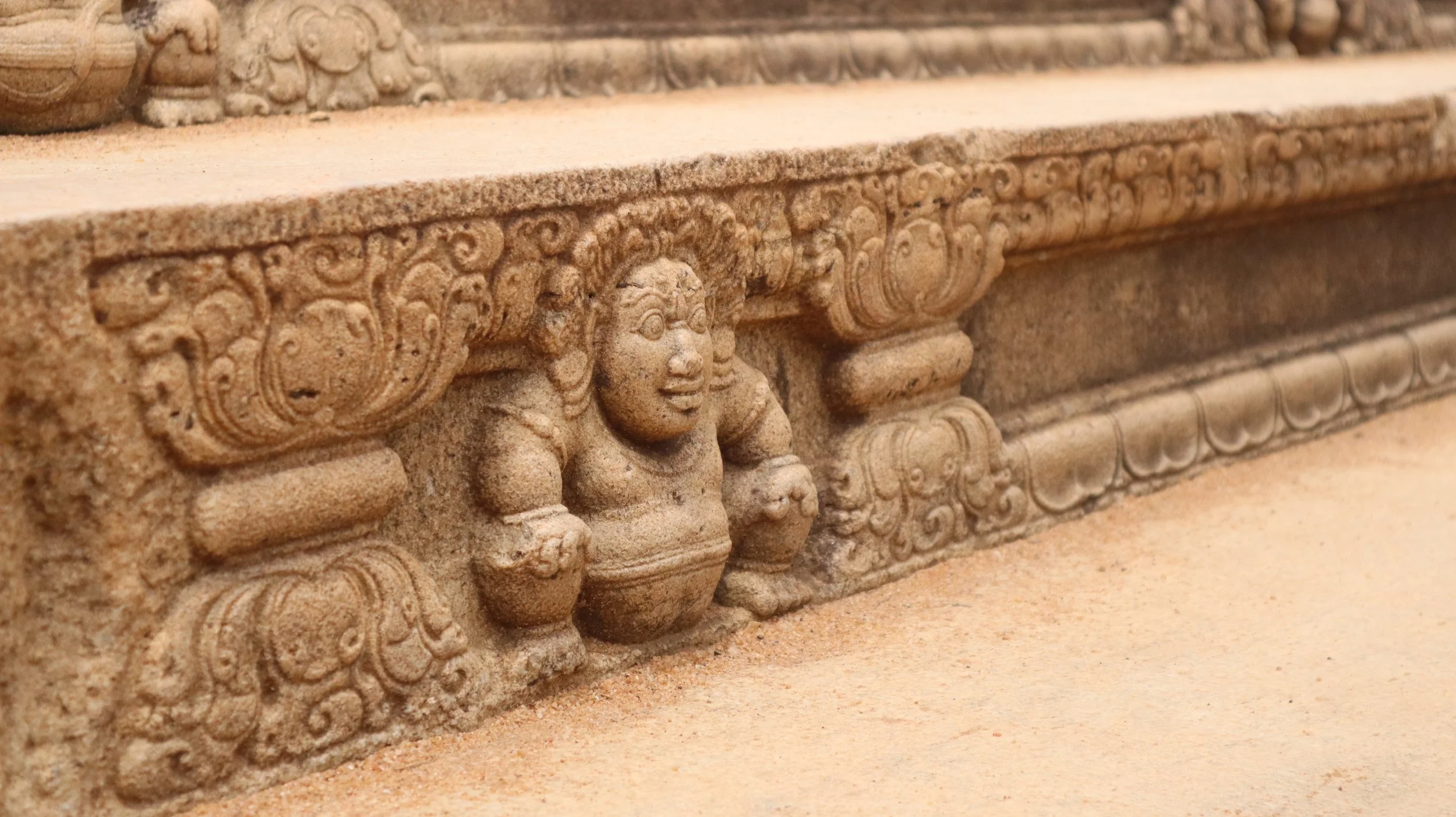Close-up of a carved sandstone decorative element featuring a smiling figure with large eyes, curly hair, and traditional attire, part of an ornate architectural frieze.