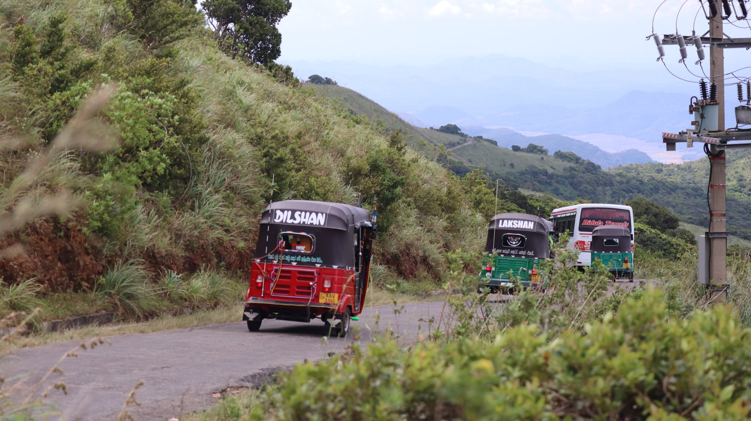 Three brightly colored auto rickshaws traveling on a hilly road surrounded by green vegetation and mountains in the background.