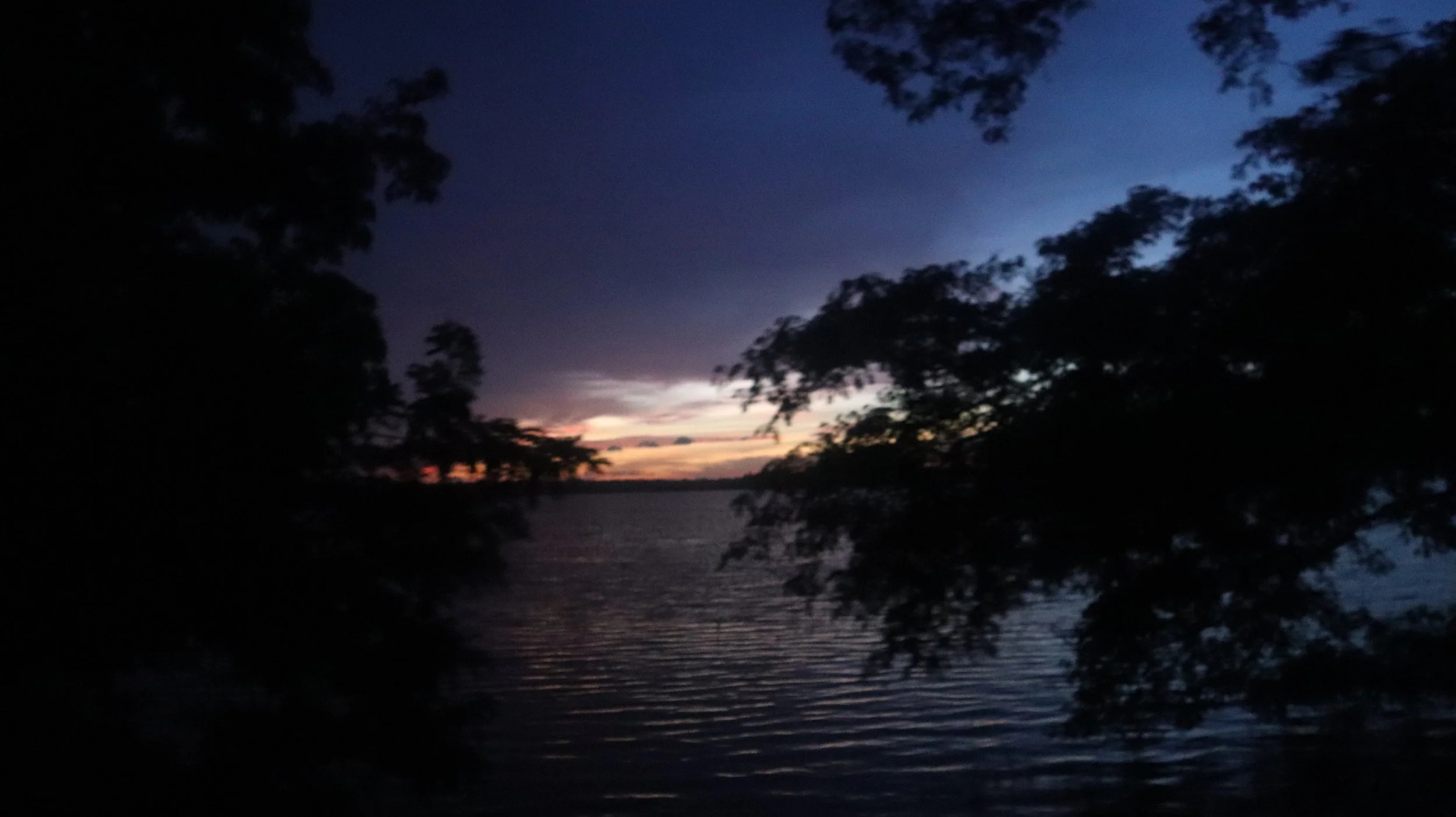 Dark lake scene during sunset with silhouetted trees on either side and a colorful sky with hints of purple, blue, and orange.