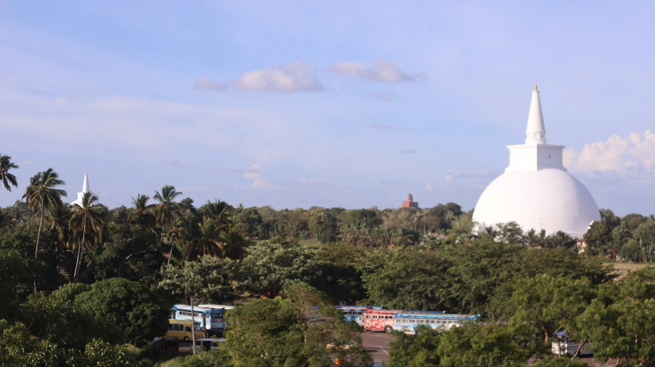 A white stupa with a spire on top, surrounded by green trees and palm trees, with a partly cloudy sky in the background.