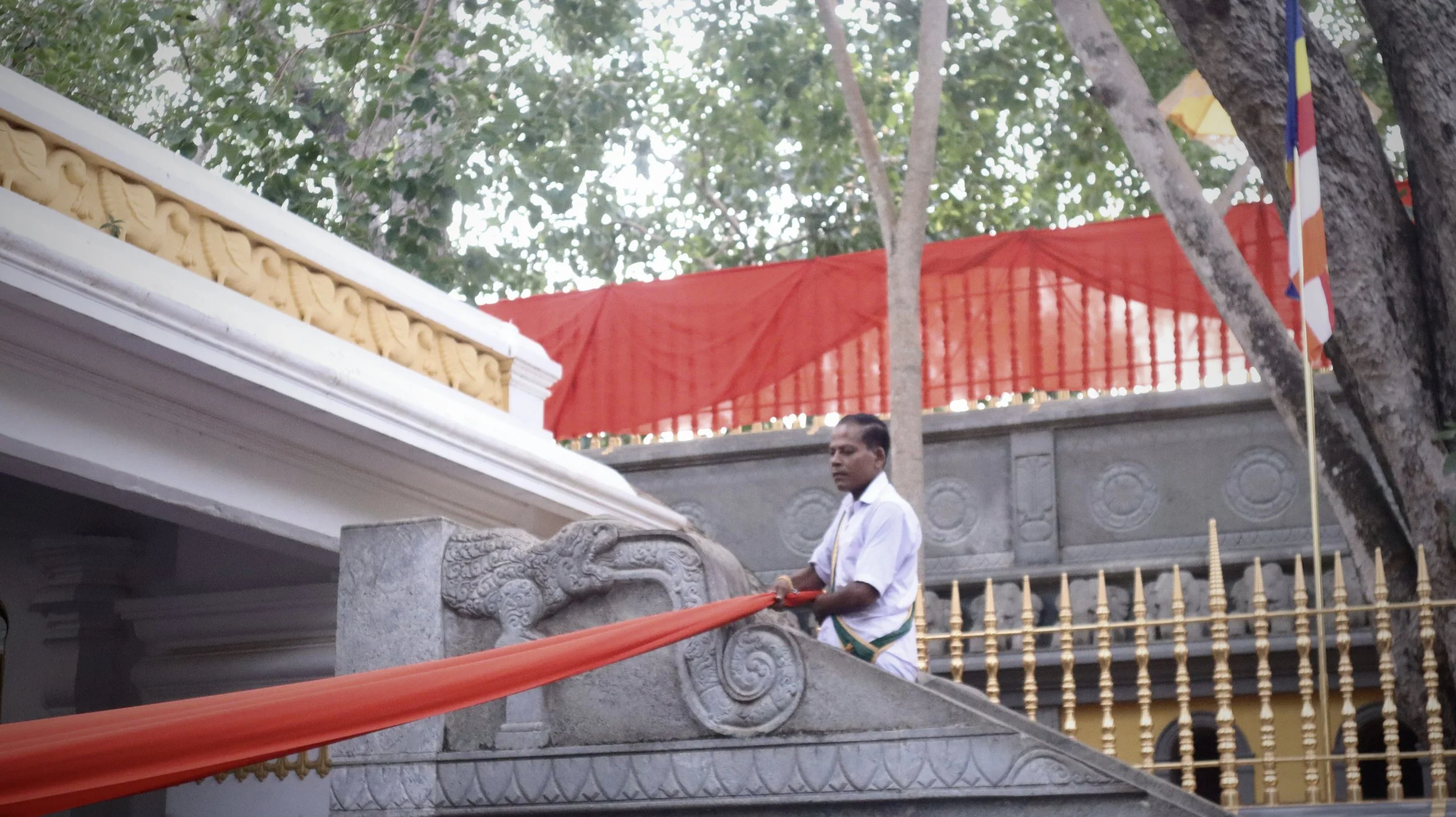 Man dressed in white wrapping orange cloth around a carved stone dragon sculpture at a temple or cultural site, with trees, a red canopy, and gold railing in the background.