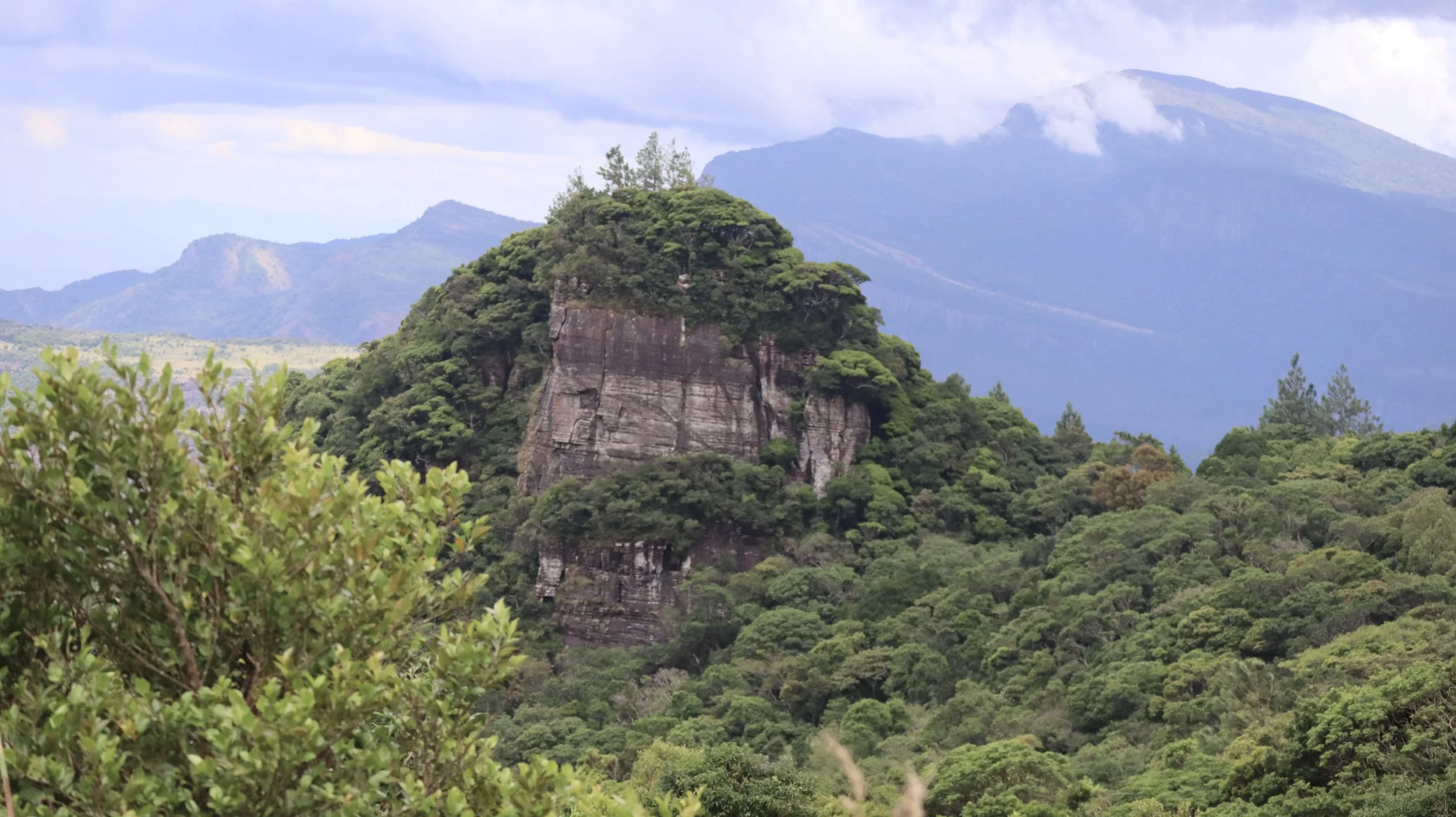 Lush green mountains and dense forest with a prominent rocky hill in the foreground, and a hazy mountain range in the background under a partly cloudy sky.