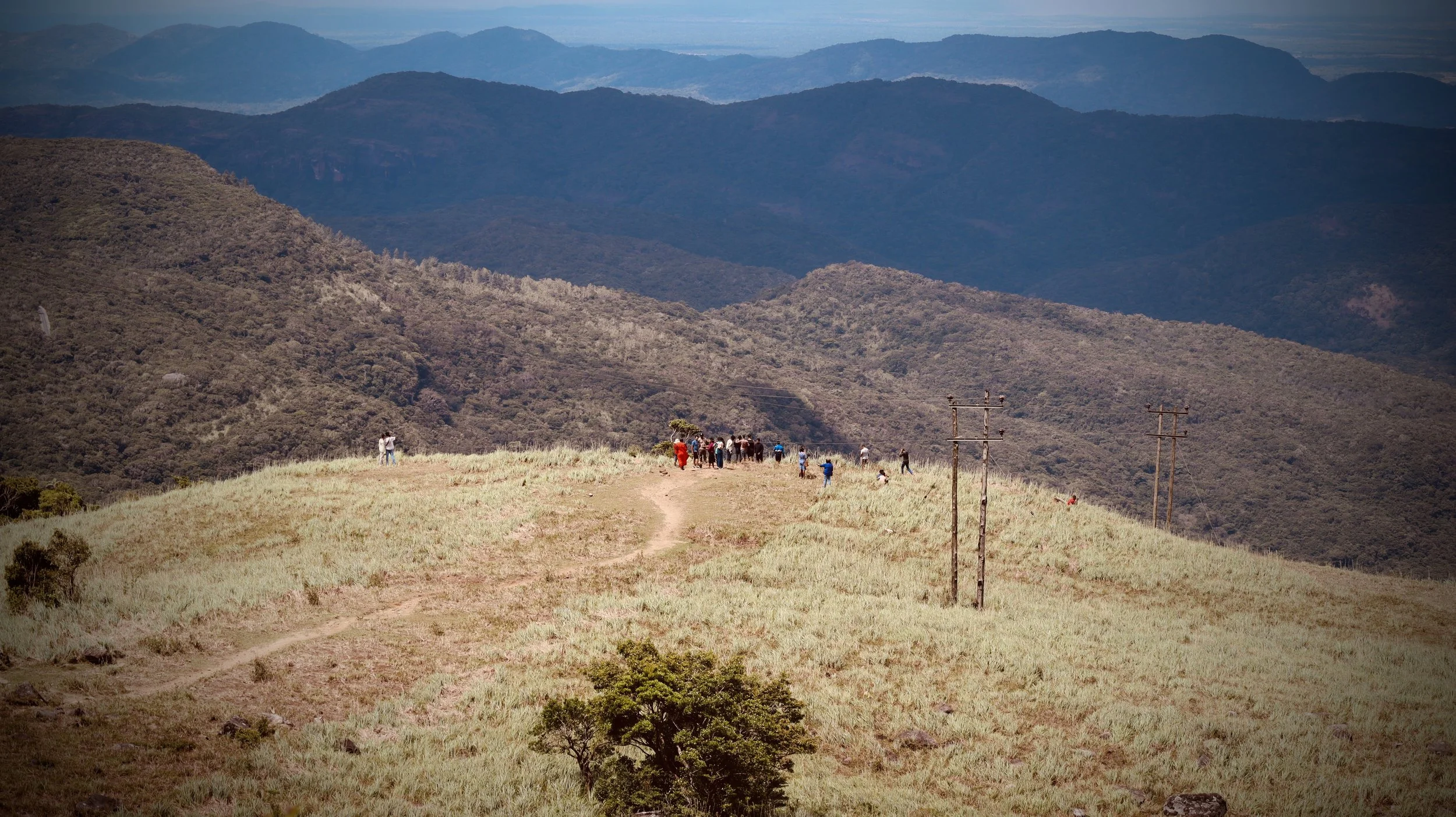 A landscape view of rolling hills and mountains with a group of people walking along a dirt trail on the grassy hillside, and power lines running across the scene.