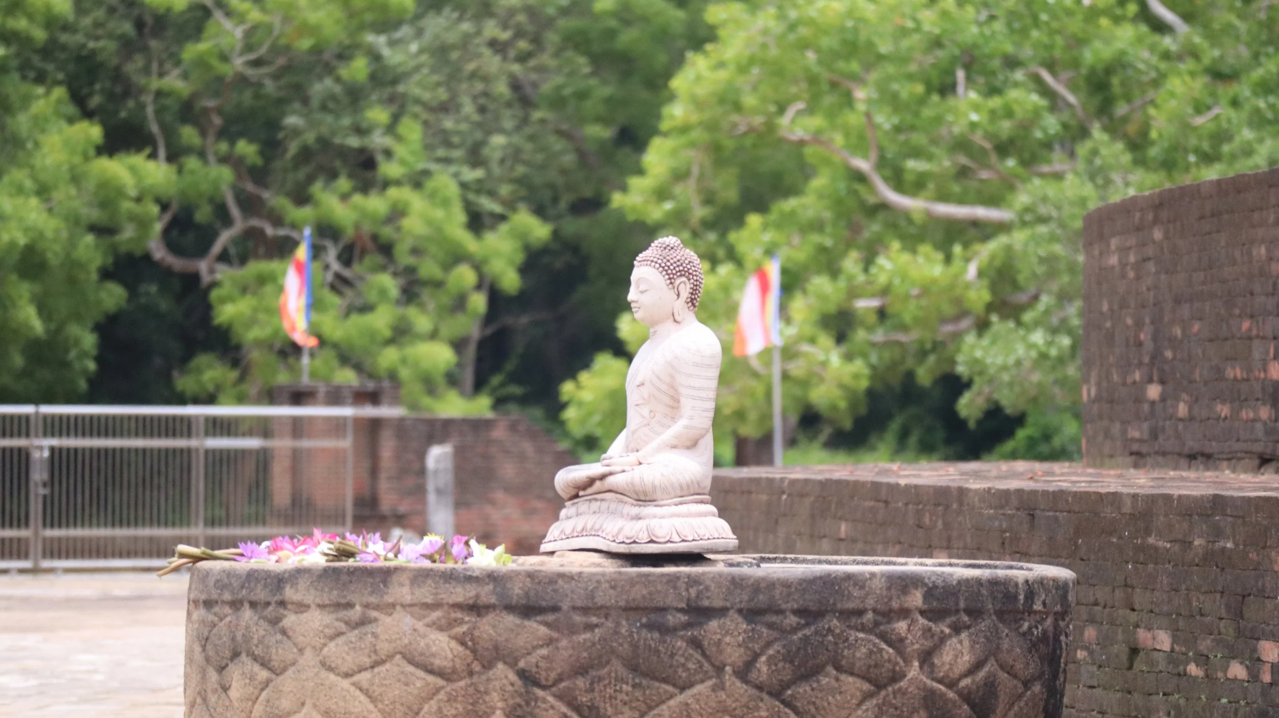 A carved white Buddha statue seated in a meditative pose on a lotus base, surrounded by pink and white flowers, with green trees and flags in the background.