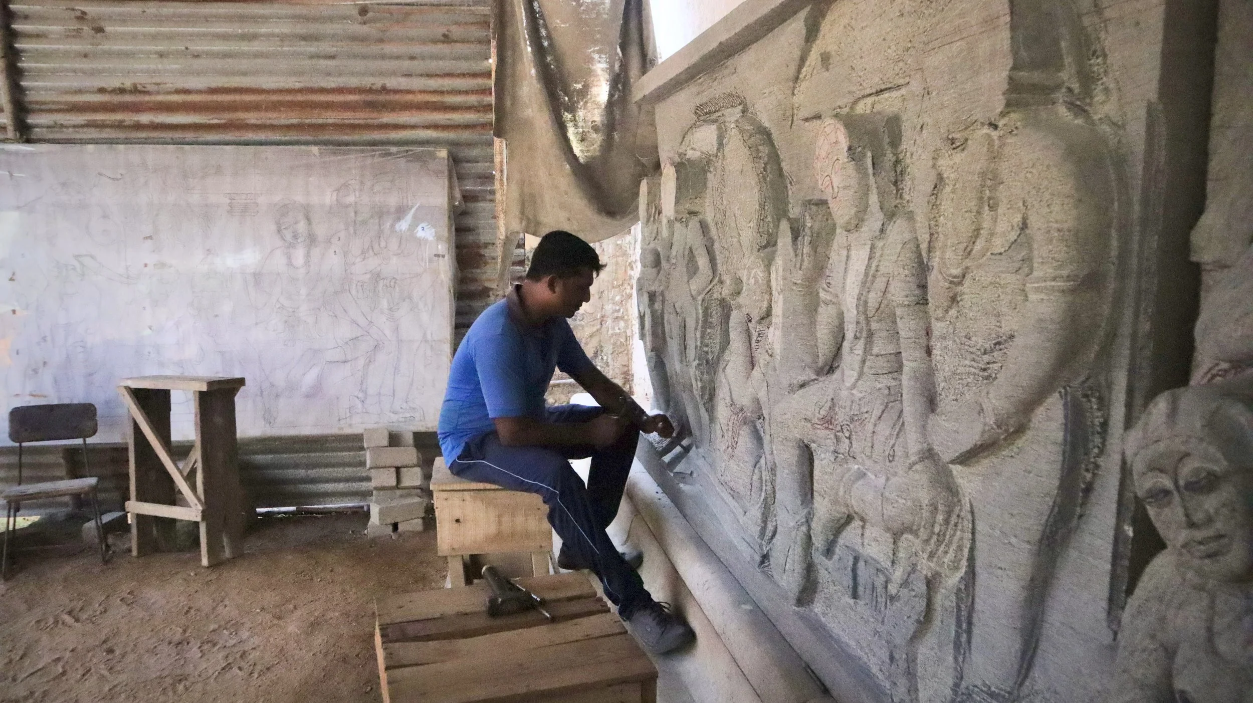 A person sculpting and carving ancient stone reliefs depicting human figures and faces inside a workshop or studio with rustic walls.