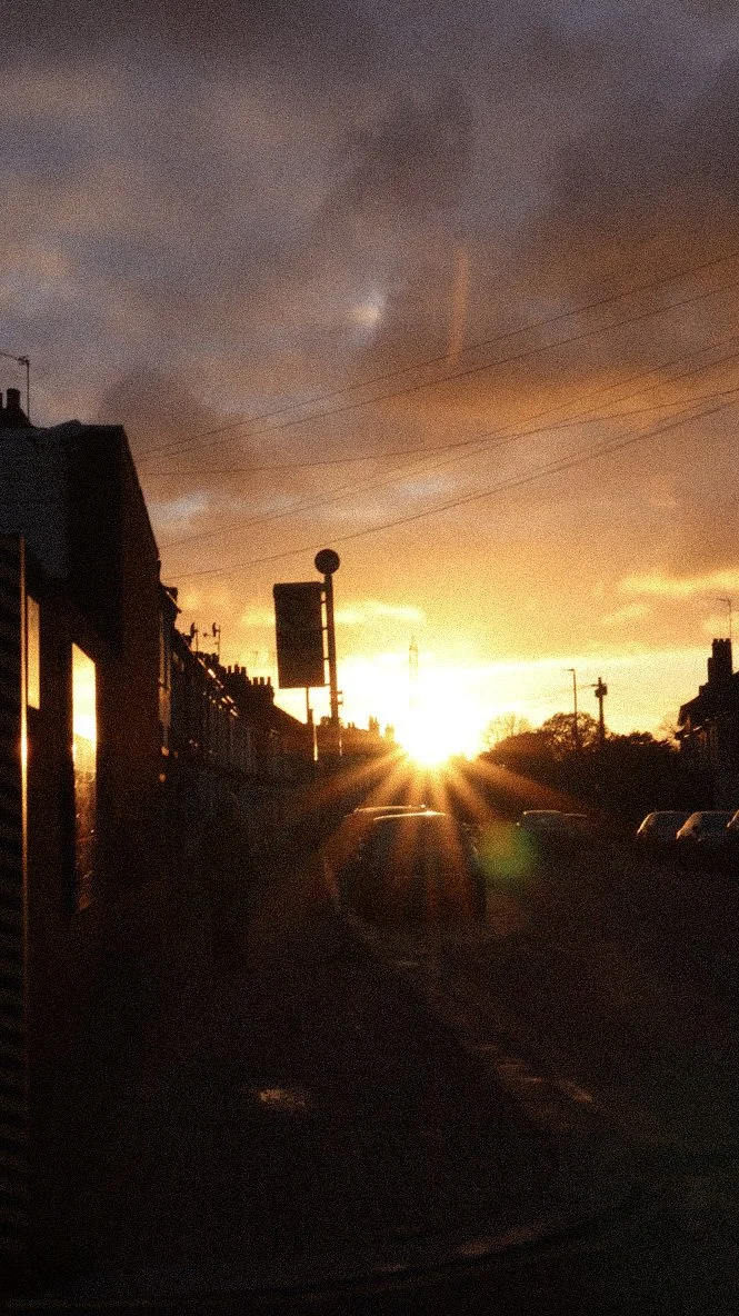 Sunset over a street with buildings and parked cars, with the sun reflecting off the windows and casting shadows.