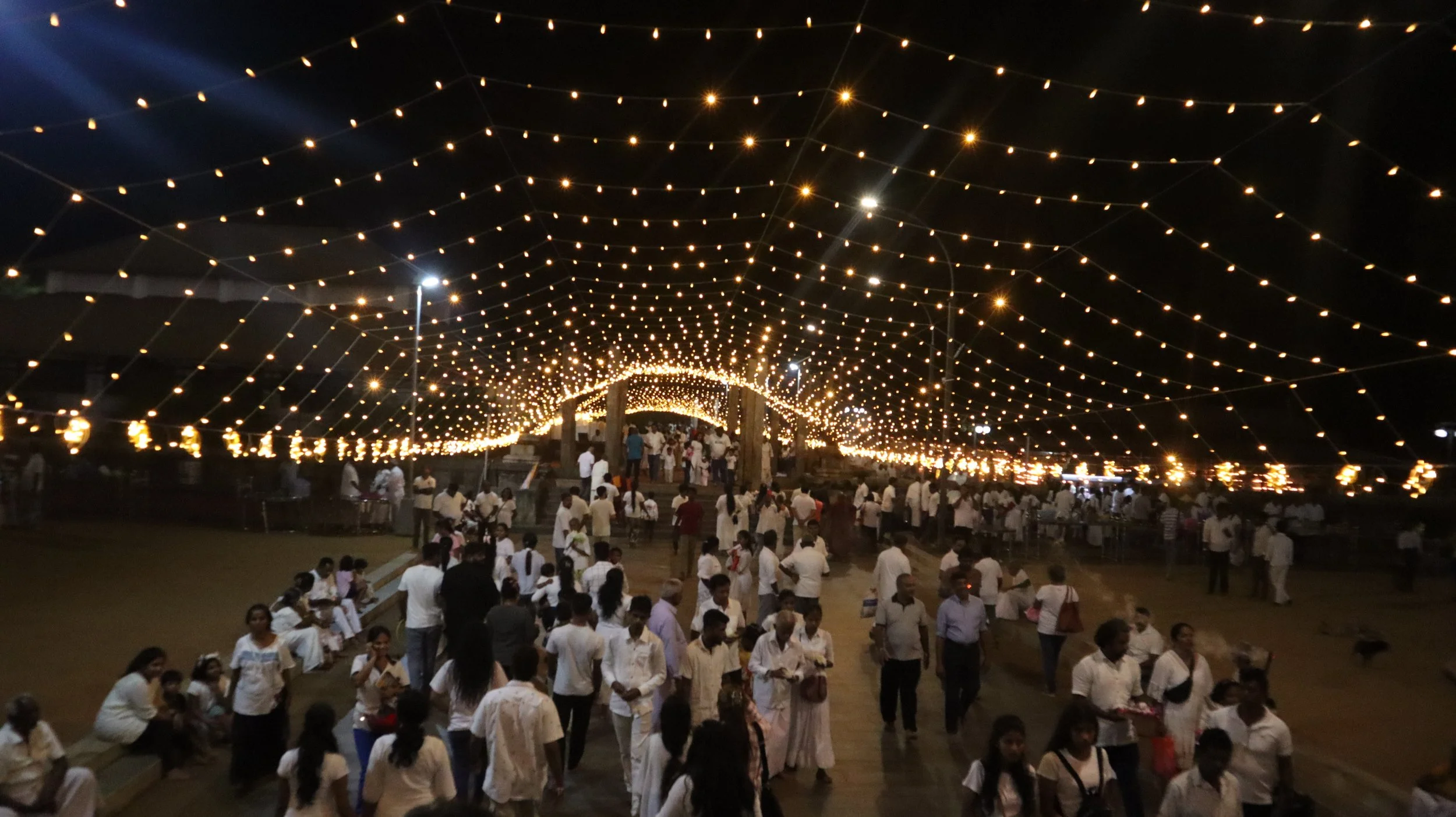 Night scene of a large crowd under a canopy of hanging string lights, with people gathered and walking around, some sitting on benches, during an outdoor event or festival.