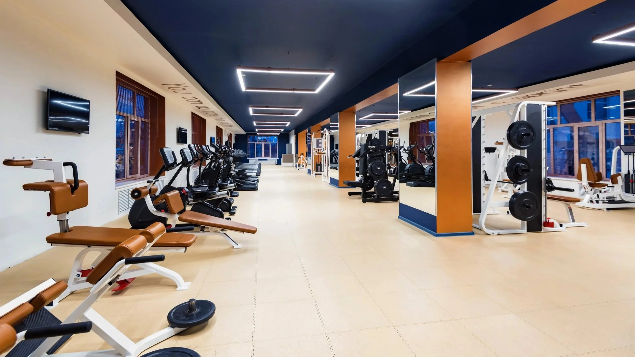 matterpot scan photo of Empty modern gym with cardio machines on the left, weightlifting equipment on the right, beige flooring, dark ceiling with rectangular lights, large windows with blinds, and a door at the back.