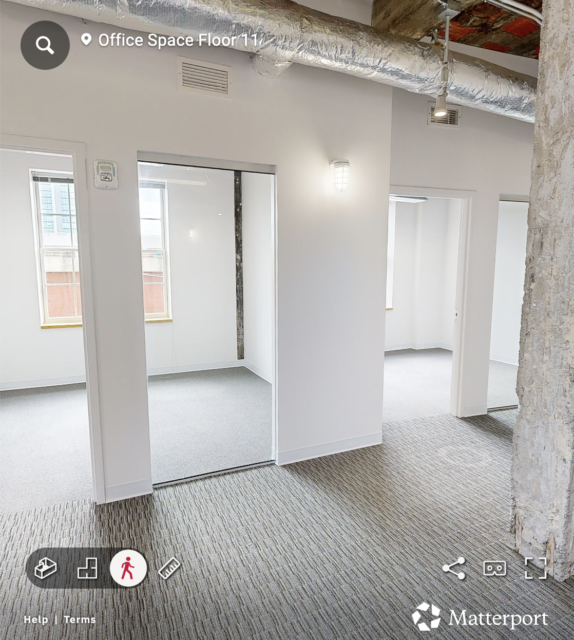 A matterport scan photo of An office space under construction with white painted walls, exposed ductwork and ceiling beams, and new carpeting. There are multiple doorways leading to small rooms, with windows providing natural light.