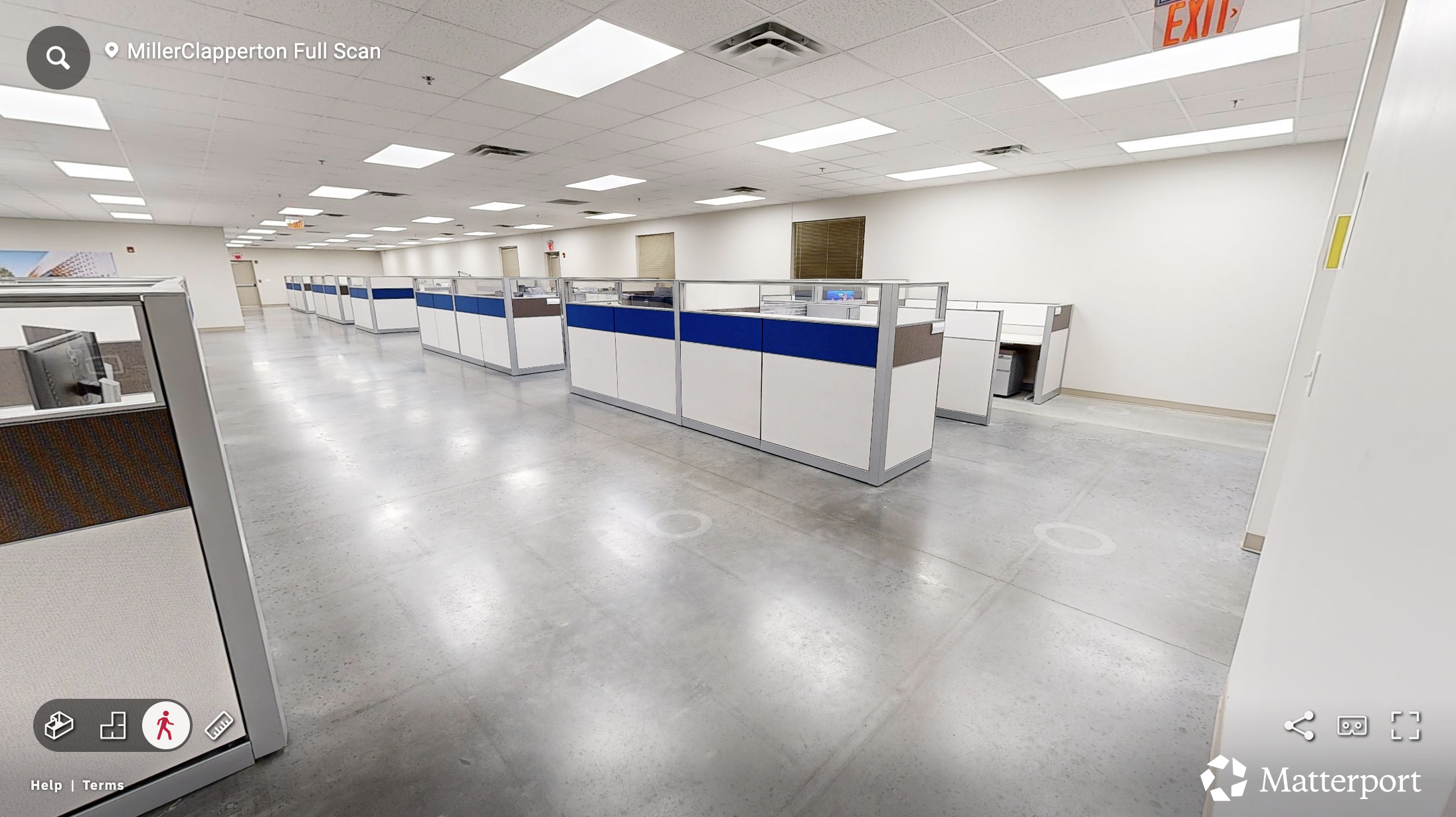 Matterport scan photo of Empty office cubicles with gray flooring, white walls, and ceiling, some computer monitors, and doors in the background.