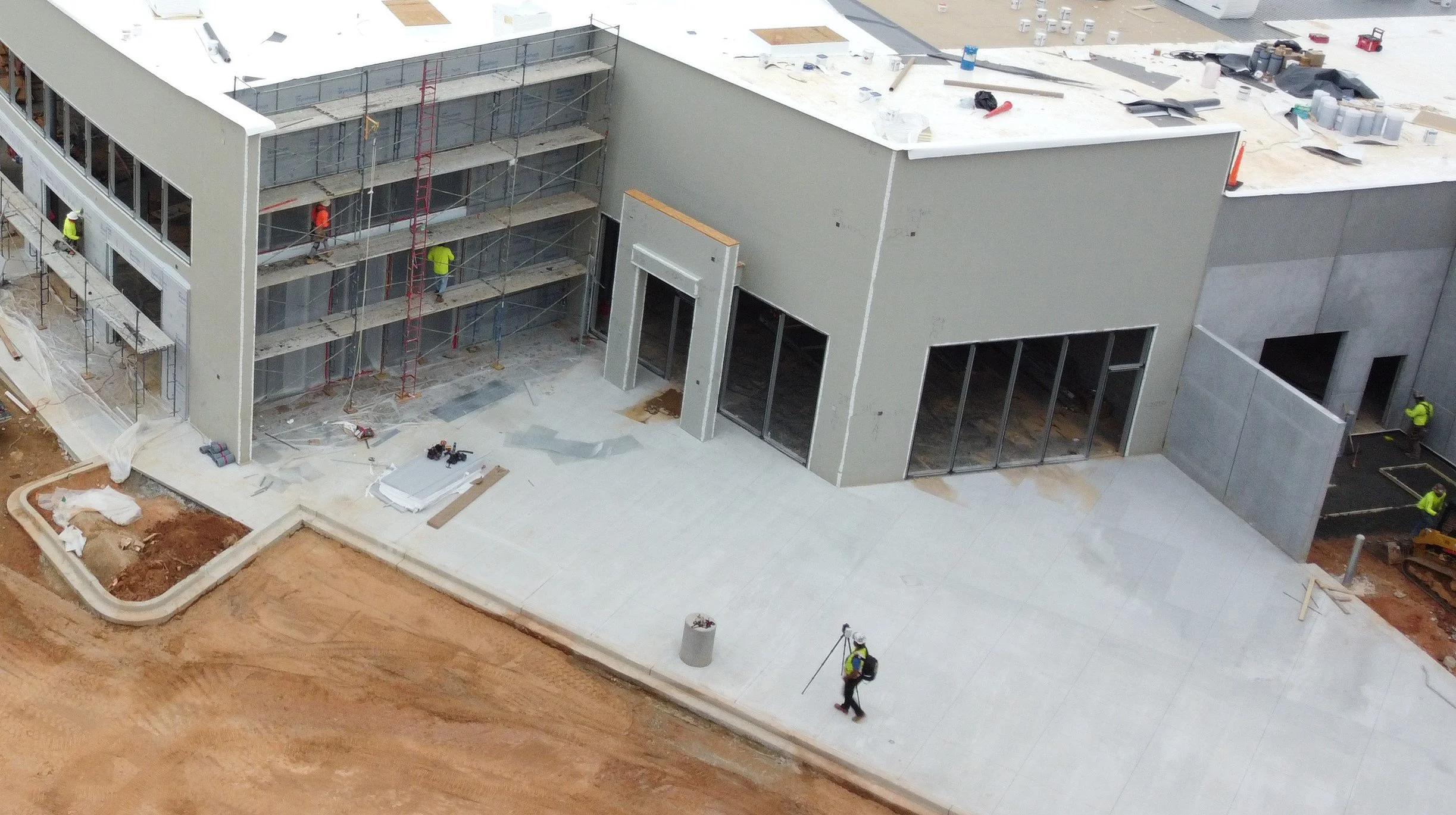 Construction workers on scaffolding and ground surrounding a modern commercial building under construction with large glass windows and exterior walls, some workers are wearing safety vests and helmets. A man walking with a laser scanning device.