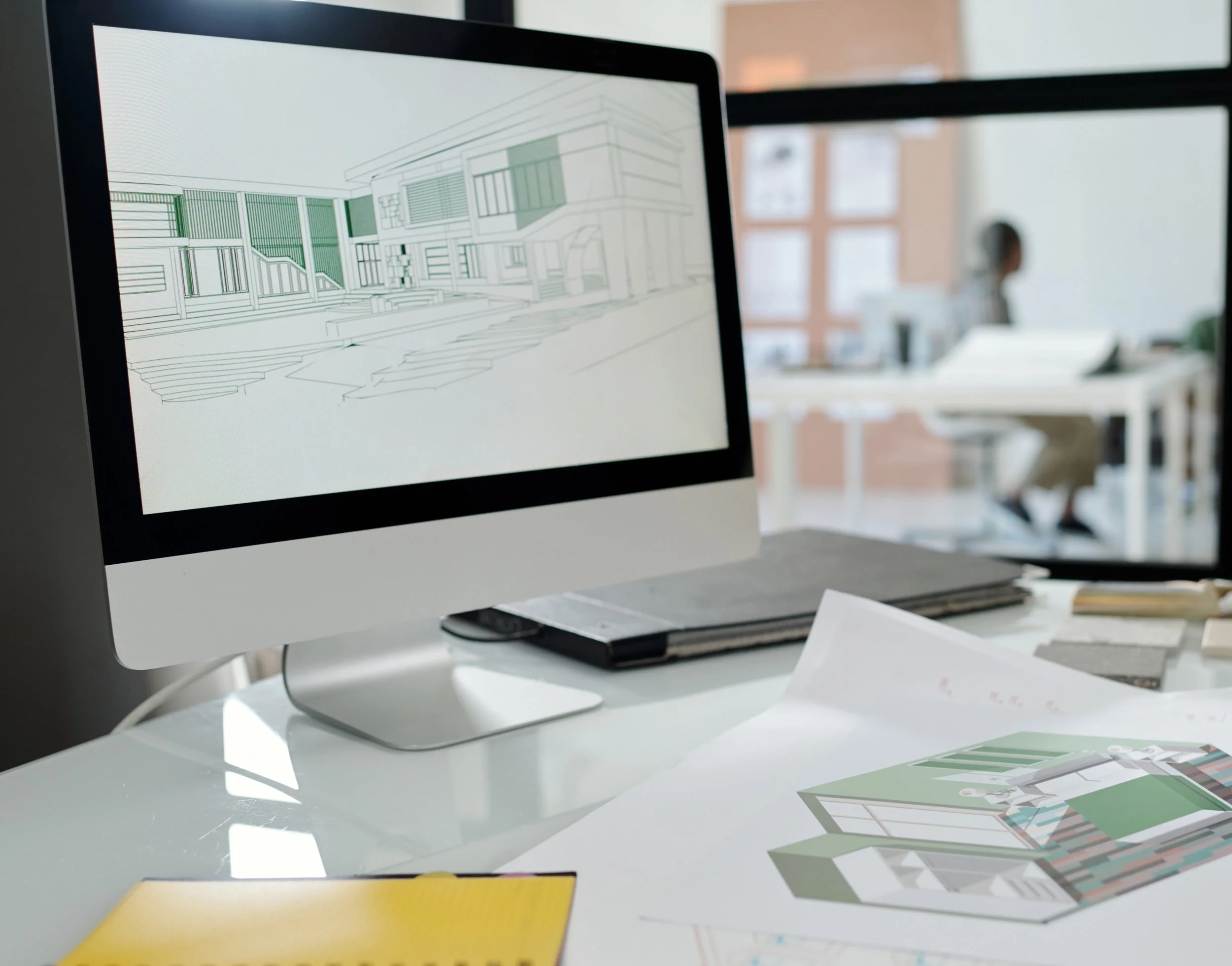 Computer monitor displaying 3d architectural sketches and design plans, with papers and color swatches on the desk.