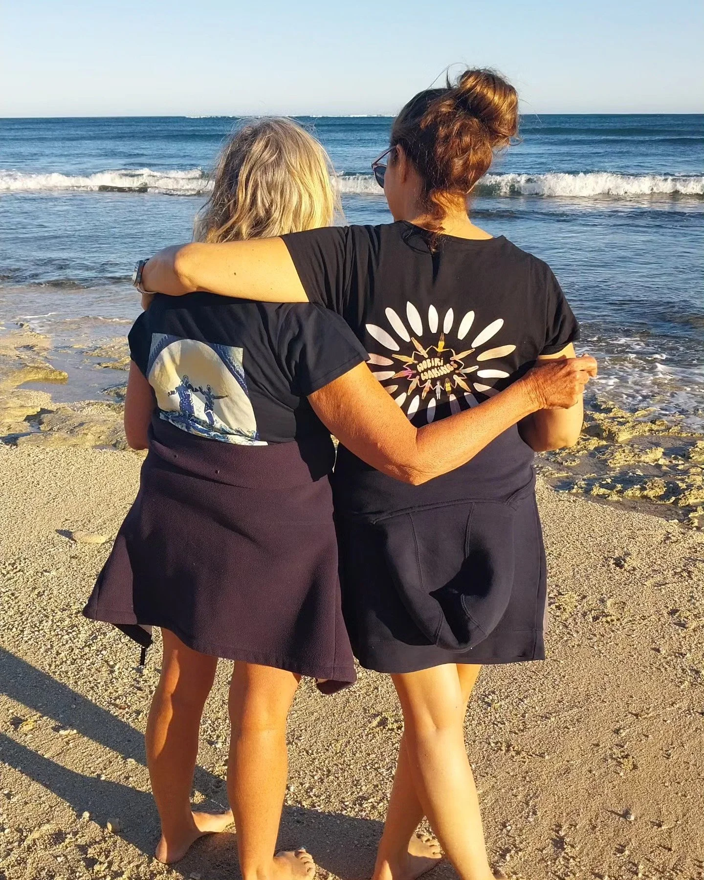Two women with their arms around each other hugging on a sandy beach, with the ocean and waves in the background, custom design for wobiri wahines