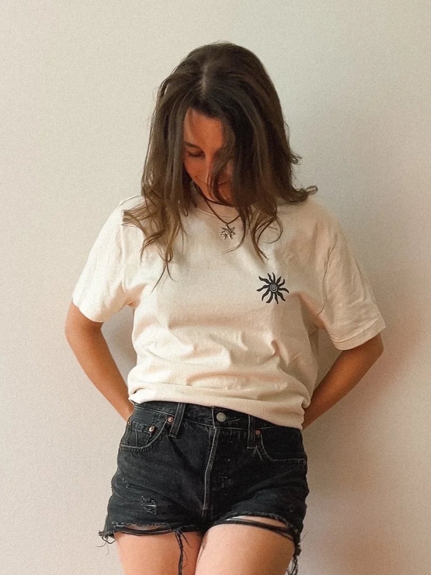 Woman with brown wavy hair wearing a white t-shirt with a sun design and black distressed denim shorts standing against a plain light-colored wall.