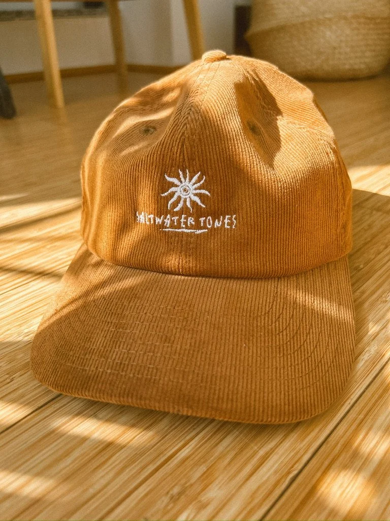 A brown corduroy cap with white embroidered logo and text reading 'Saltwater Tones' on the front, placed on a wooden surface.