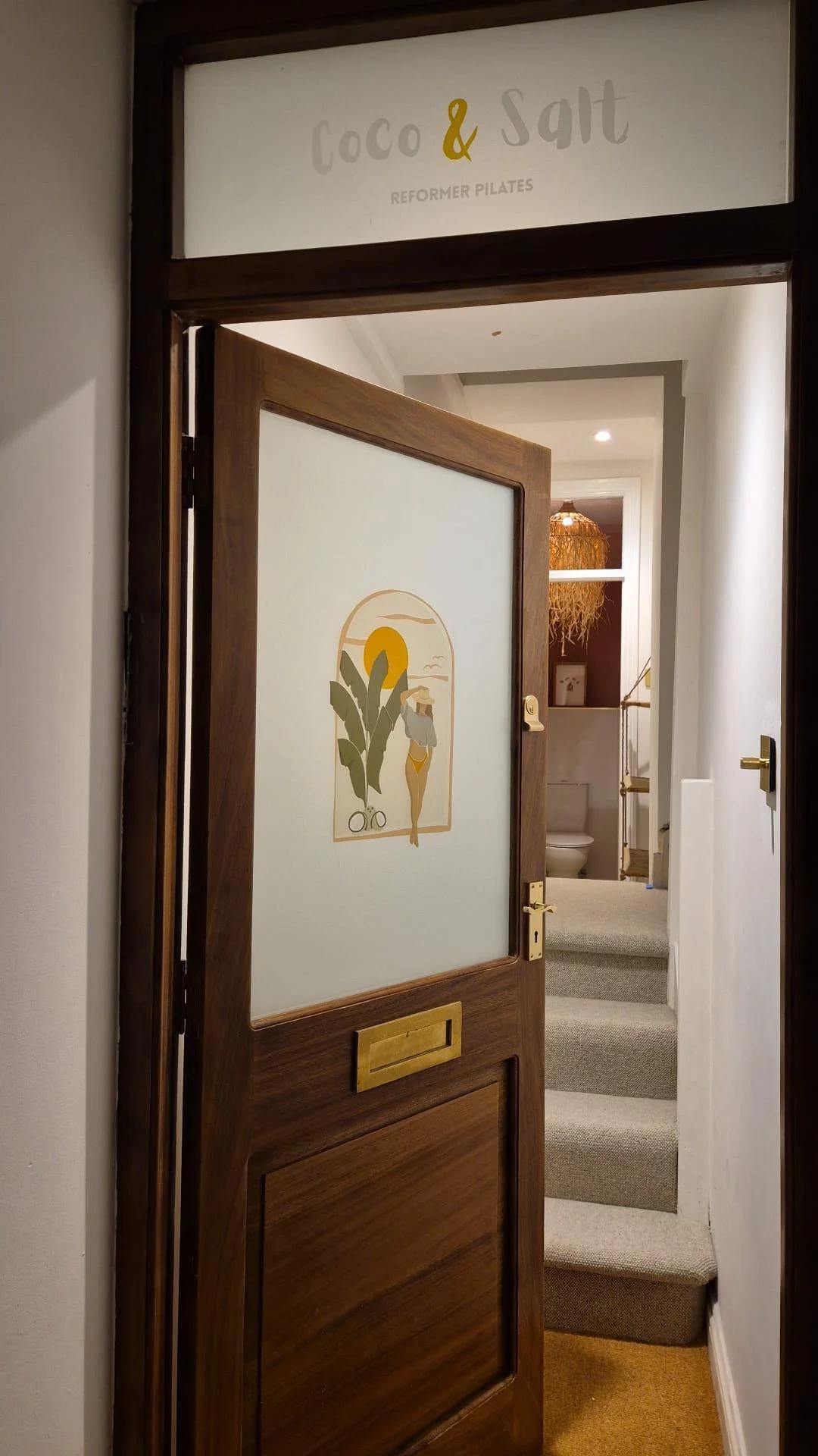 Interior view of a bathroom entrance with a wooden door featuring a frosted glass panel. The panel has an illustration of a woman in a hat with a plant, a sun, and clouds. The doorway leads to a staircase and a bathroom with a toilet, wooden shelving