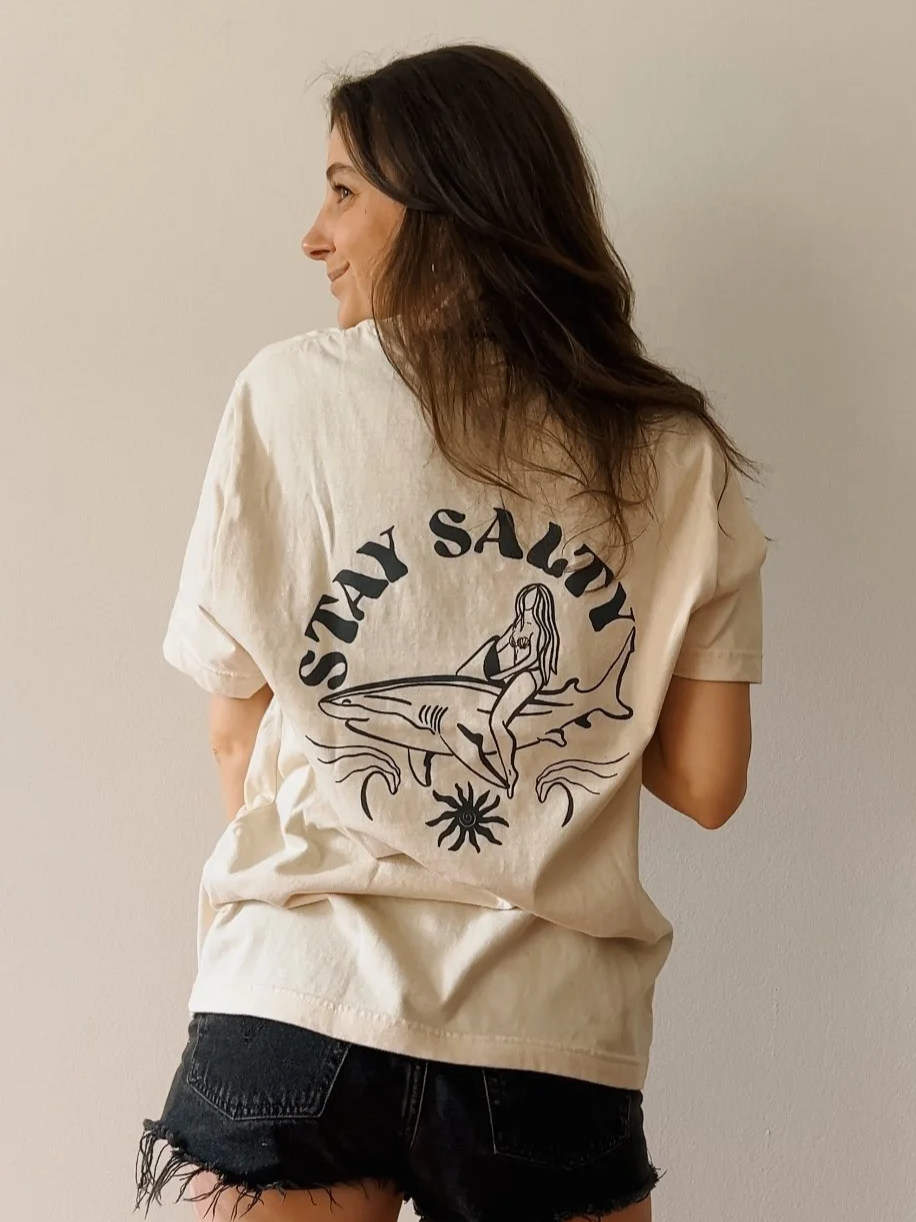 A woman with long brown hair wearing a beige T-shirt with a graphic of a woman riding a shark and the words 'Stay Salty,' paired with black ripped shorts, standing against a plain wall.
