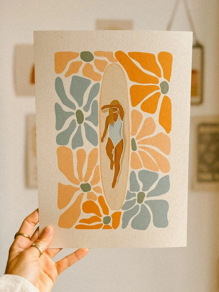A person holding up a colorful, abstract art print featuring a woman in a swimsuit and hat, surrounded by large, stylized flowers in pastel orange, blue, and green tones.