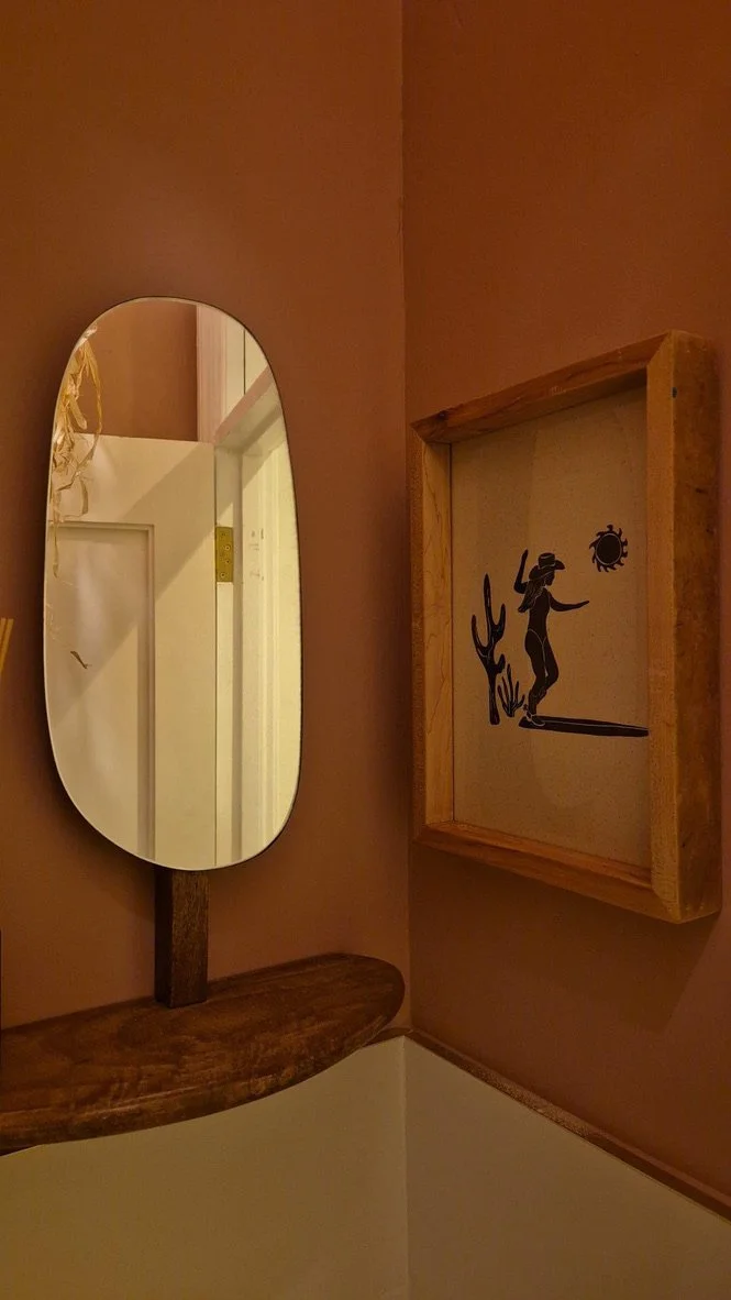 A bedroom corner with a vertical oval mirror mounted on a wooden support, a wooden shelf below it, and a framed silhouette art of a girl watering a cactus with a sun and a bug in the background on the wall.