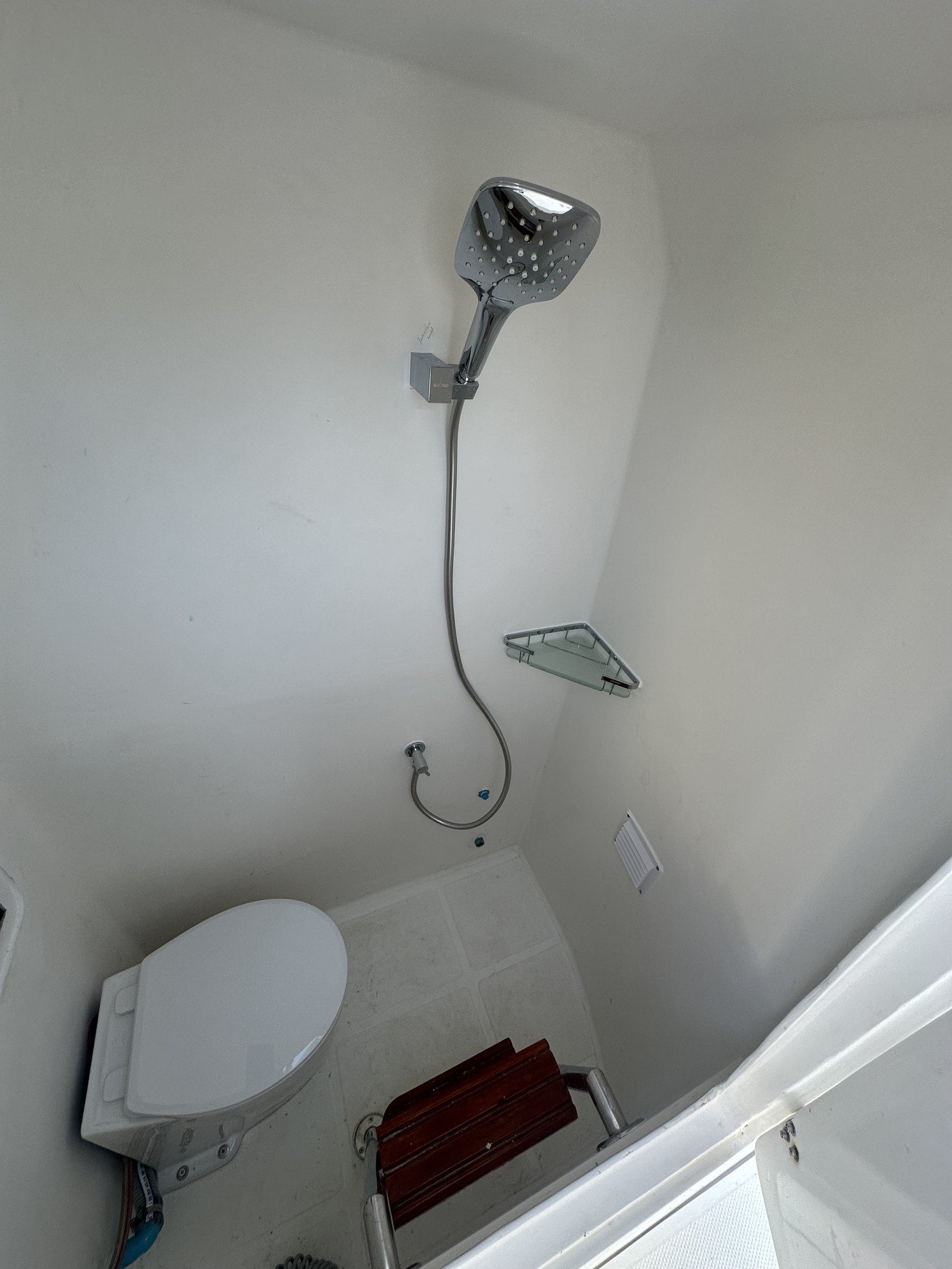 A small bathroom featuring a shower head with a hose, a mirror shelf, a white toilet, a wooden bench, a vent, and a no-slip textured floor.